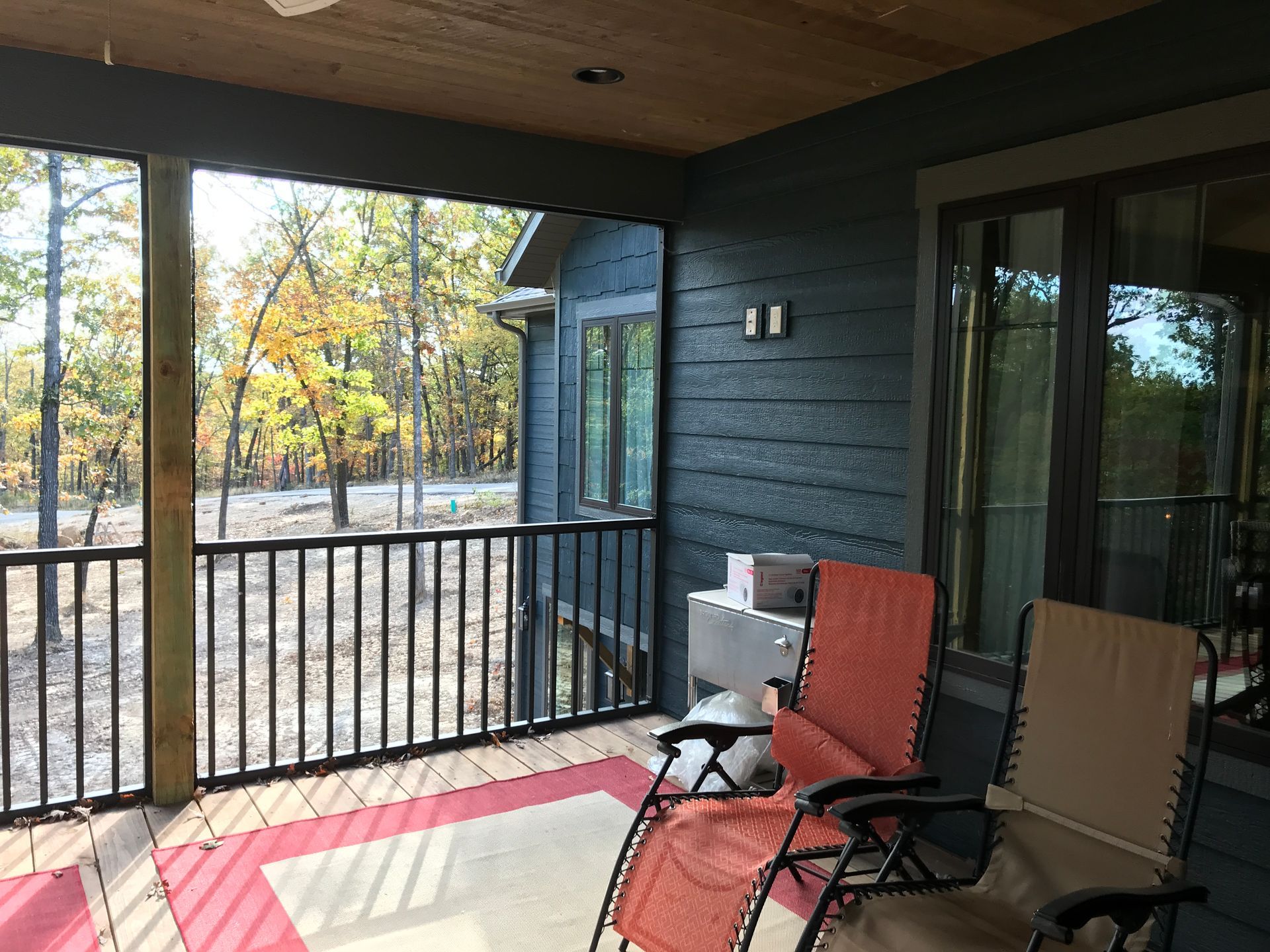 Covered porch with two lounge chairs, red rug, and a wooded view through black railings