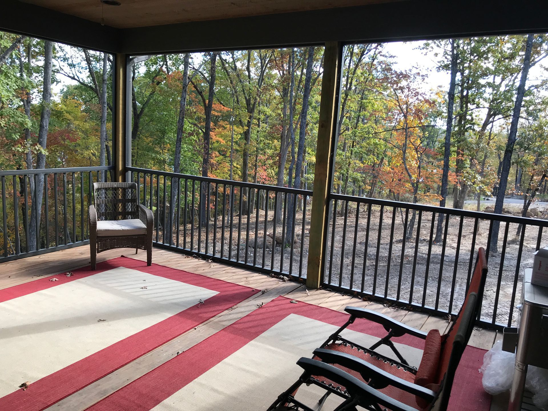 Covered porch with red and beige rug, black railing, and two chairs overlooking trees in fall.
