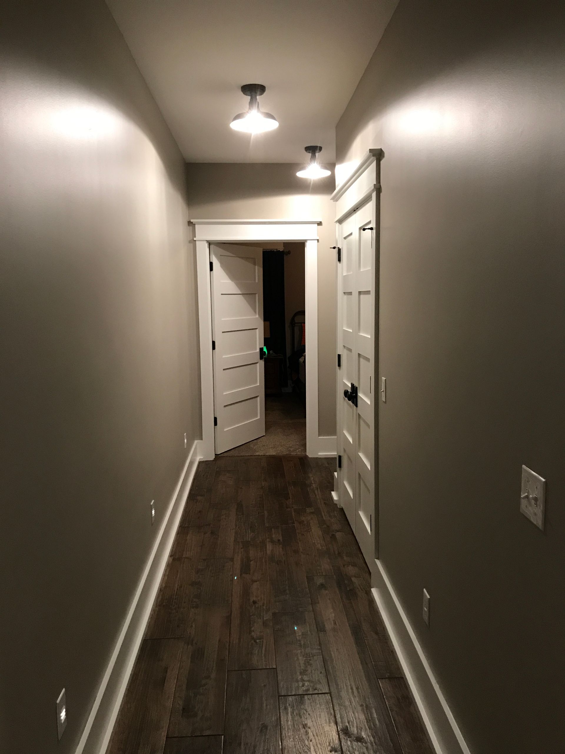 Narrow hallway with dark wood floor, beige walls, ceiling lights, and open white doors at the end.