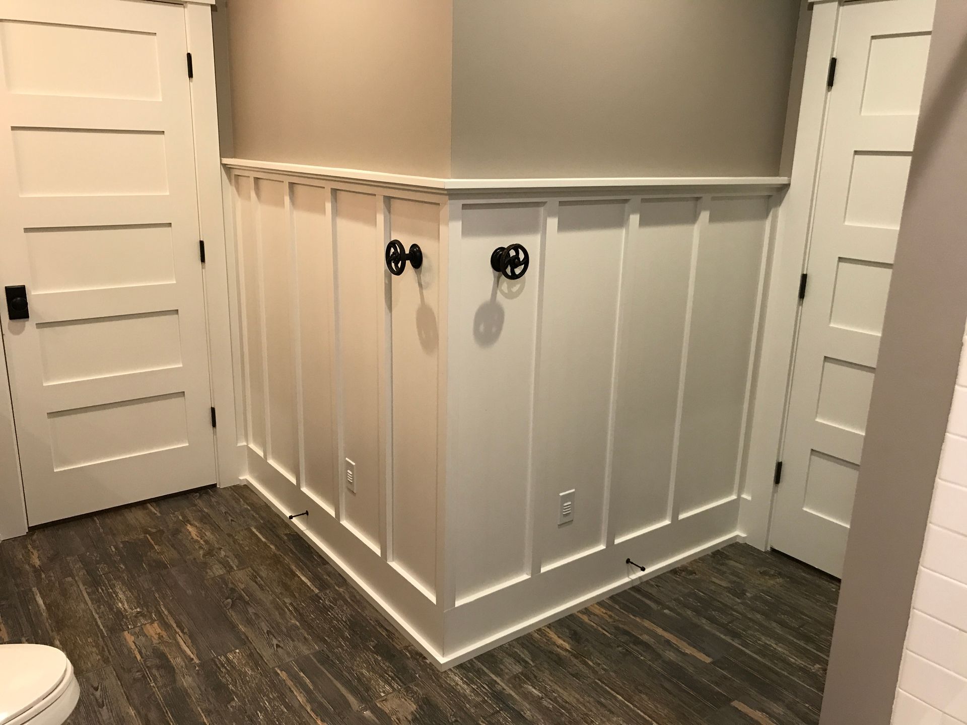Empty room with white paneled walls, dark wood floor, and two black wall hooks near a small outlet.