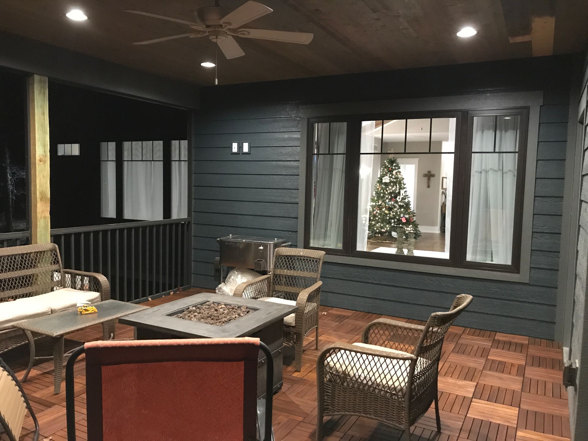 Covered patio with wicker seating, fire table, and a lit Christmas tree seen through a window