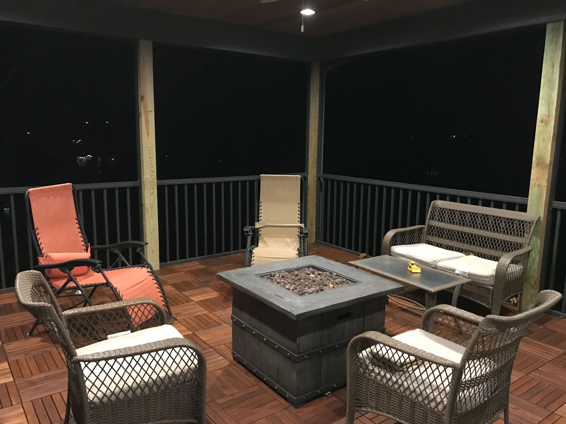 Covered deck with wicker chairs and a square fire pit at night.
