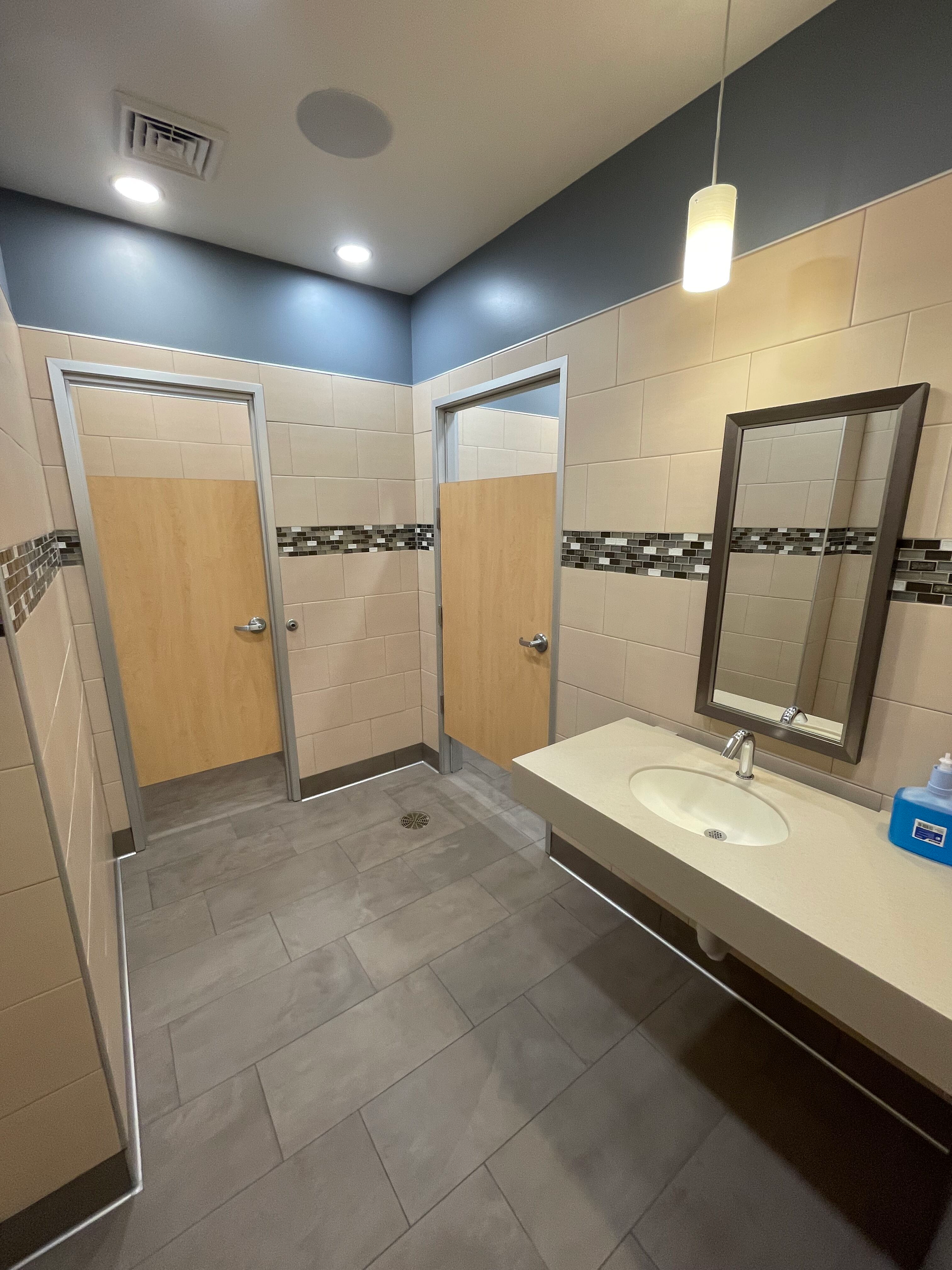 Public restroom with beige stalls, gray tile floor, and sink counter with mirror.