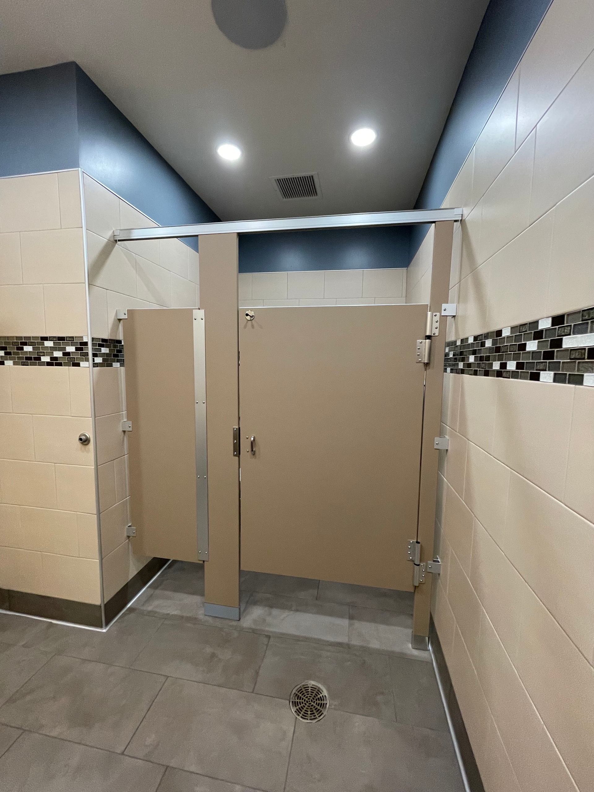 Public restroom shower stalls with beige tile, gray floor, and ceiling lights