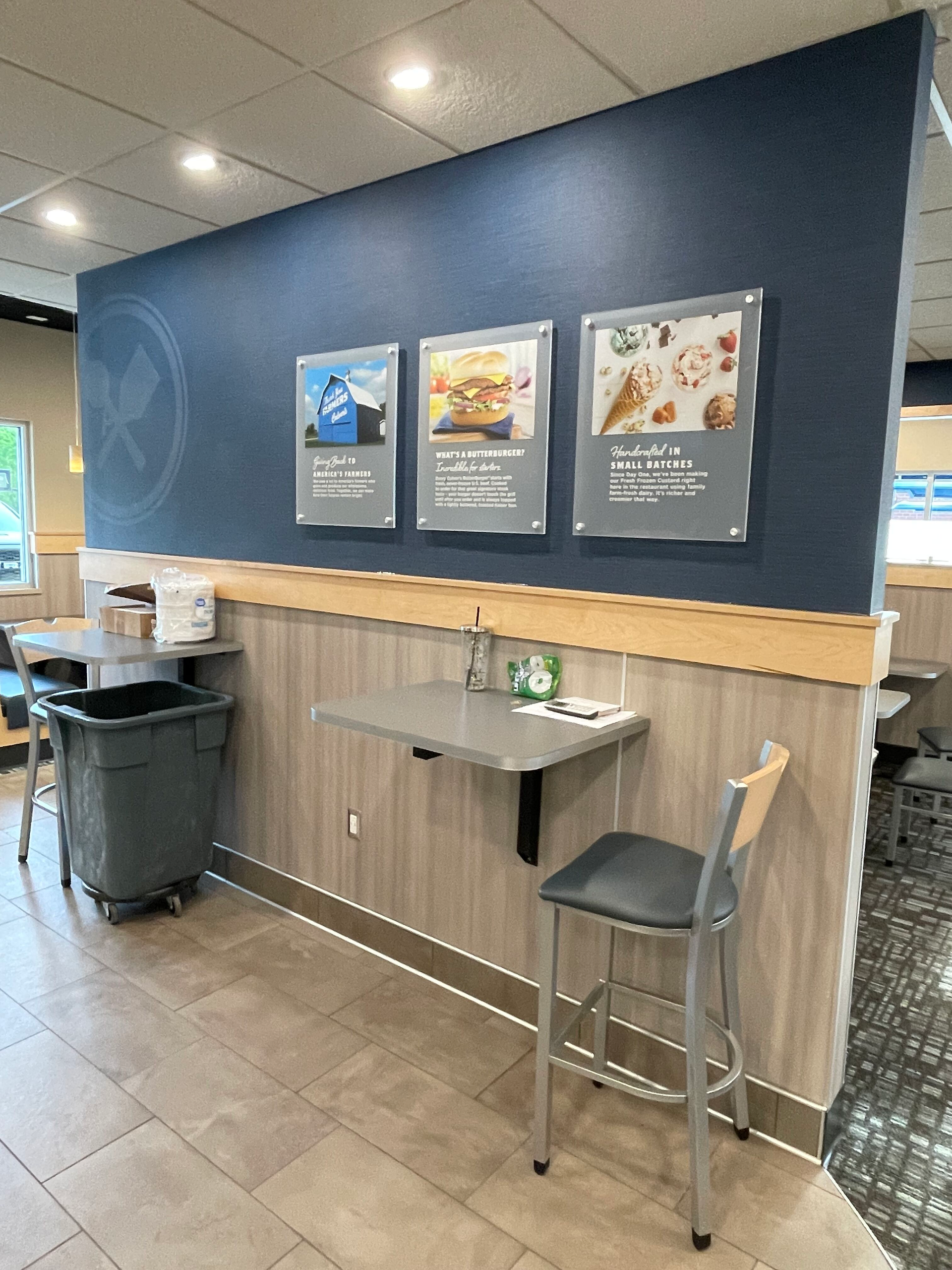 Small self-serve café counter with menu boards, a stool, and trash bin against a blue wall