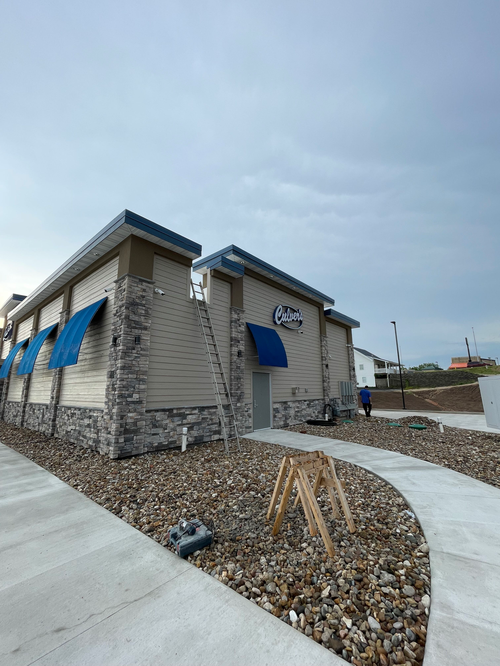 Small beige building with blue awnings, stone base, and curved concrete walkway under a cloudy sky.