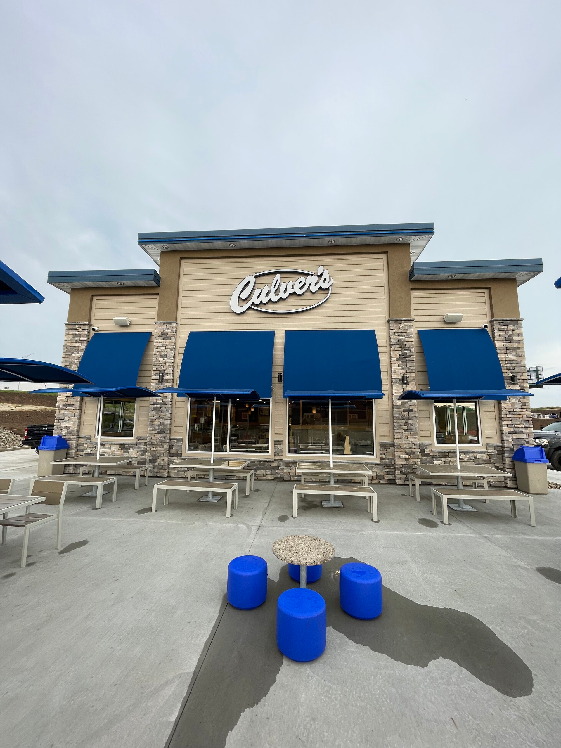 Culver’s restaurant exterior with blue awnings, outdoor tables, and blue barrels in front.