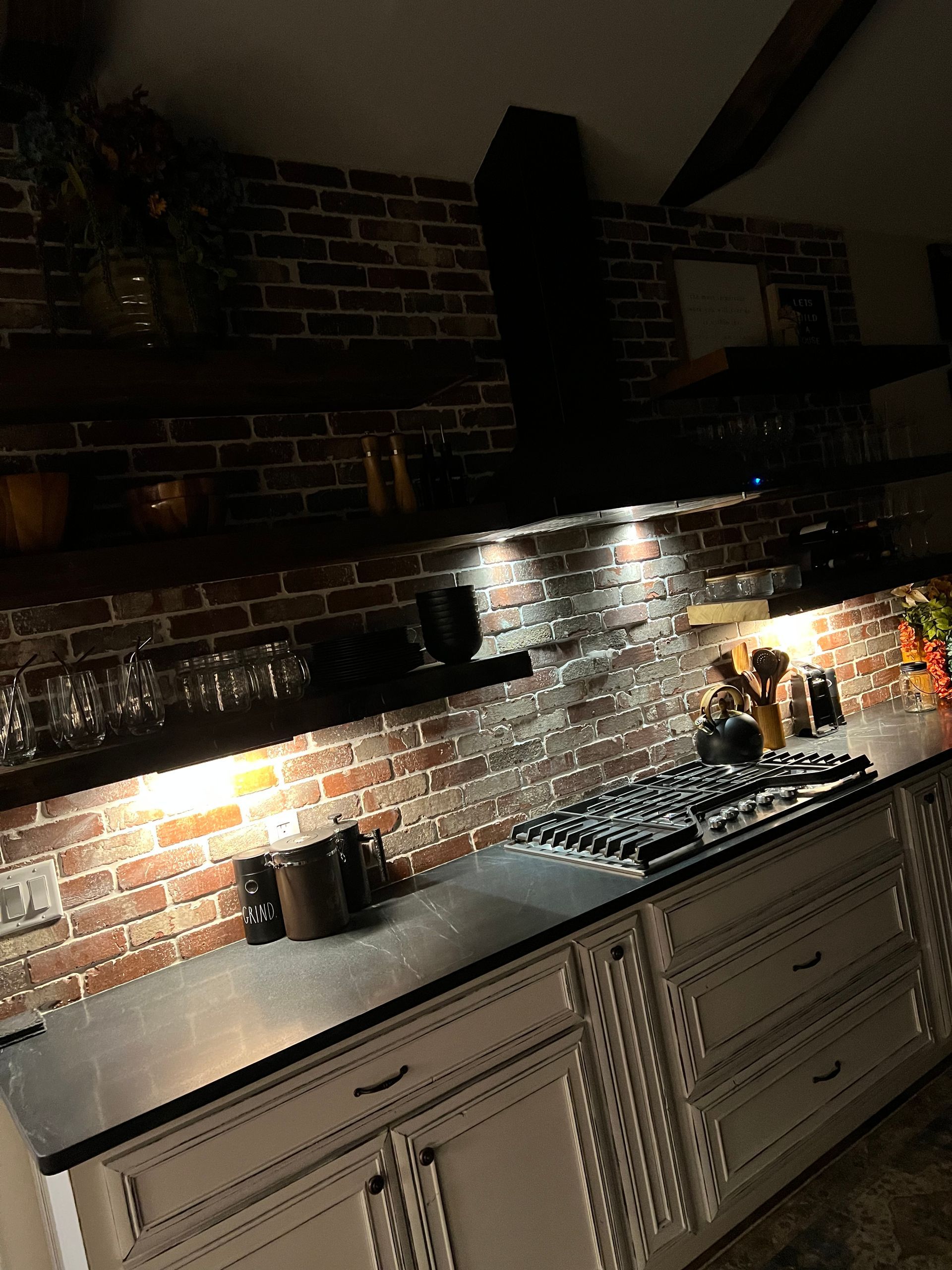 Dim kitchen with brick backsplash, open shelves, under-cabinet lighting, and a long gray countertop.