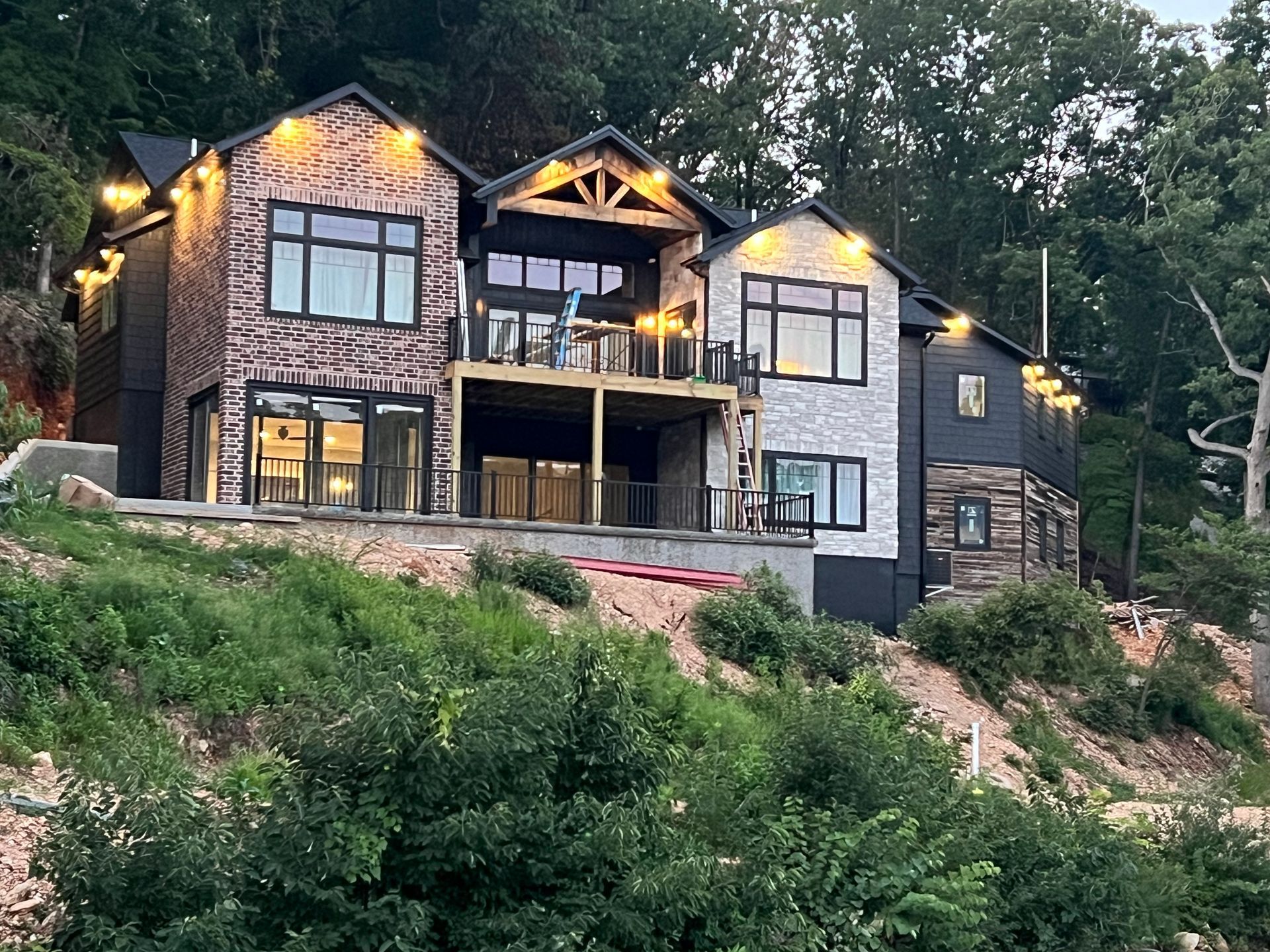 Modern stone-and-wood house on a wooded hillside at dusk, with warm exterior lights on and balconies visible