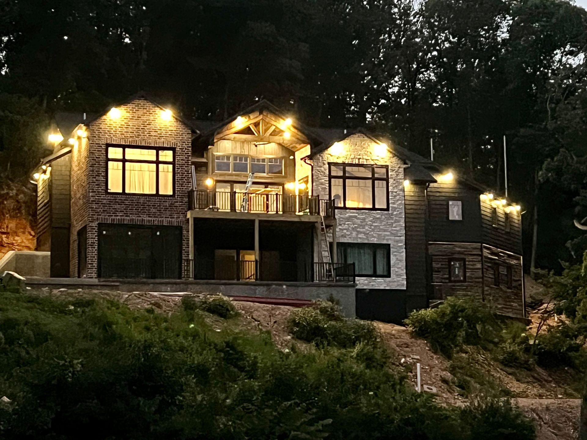 Large illuminated stone house on a wooded hillside at night, with warm lights glowing from the windows.