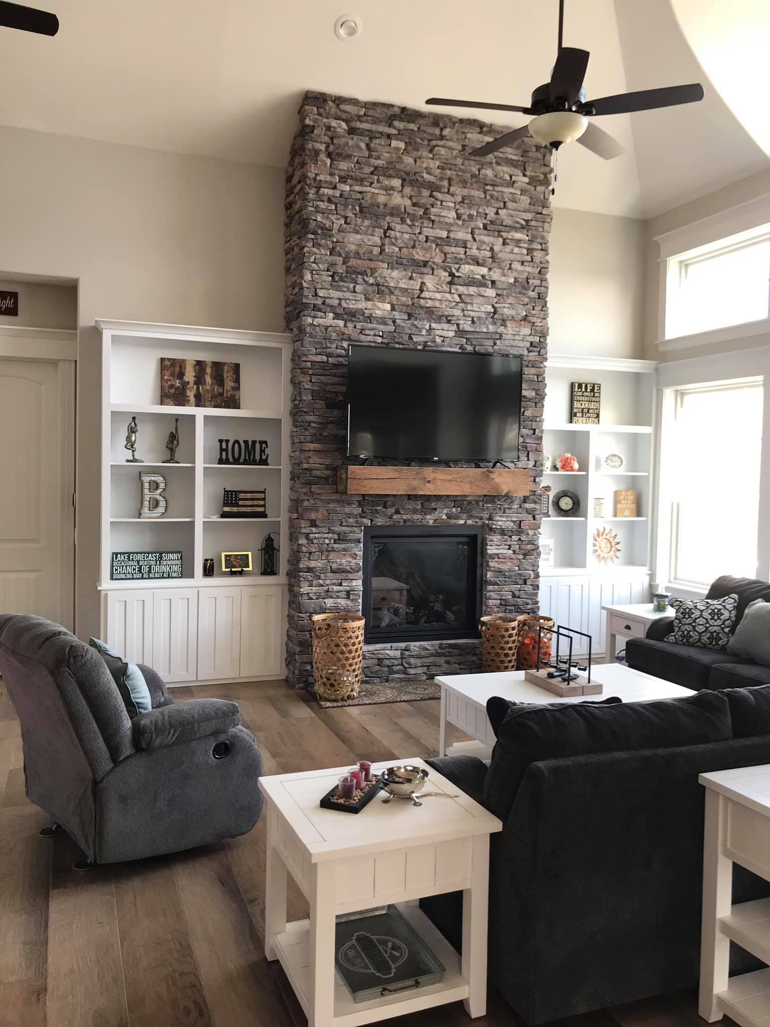 Modern living room with stone fireplace, TV, white shelving, gray and black sofas, and bright windows