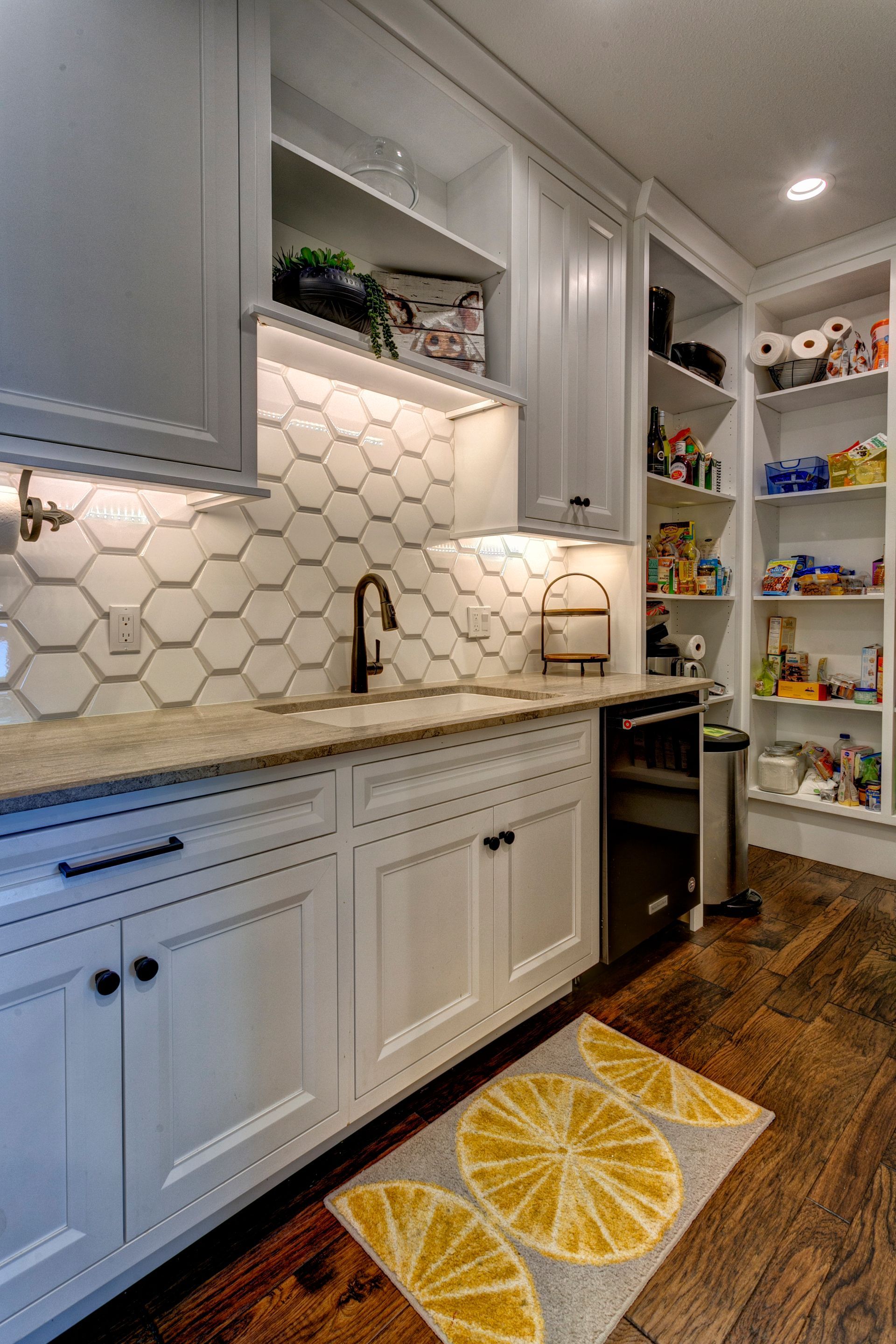 Bright kitchen with white cabinets, hex tile backsplash, open pantry shelves, and lemon-patterned rug.