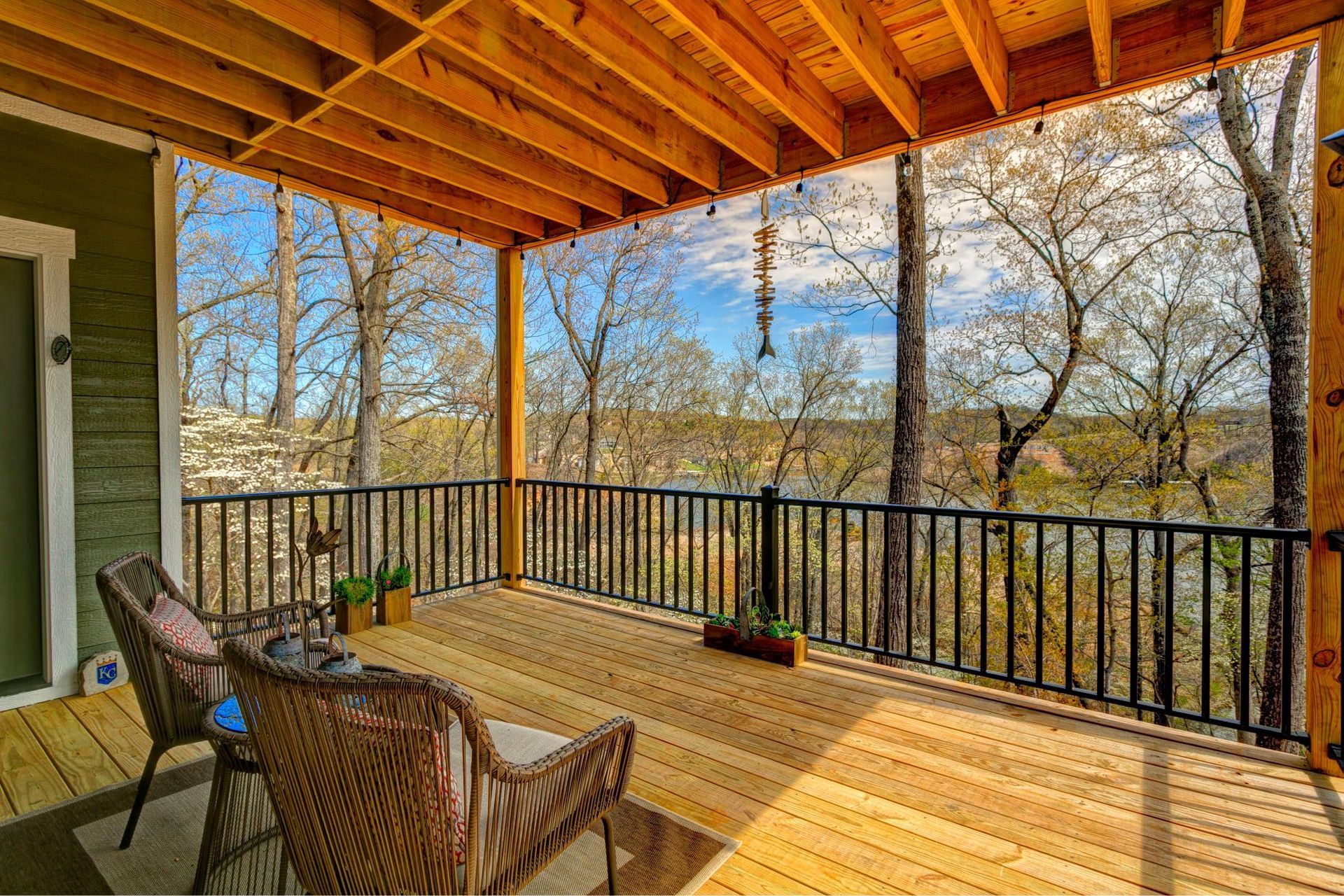 Covered wooden deck with wicker chairs, black railing, and a wooded view beyond the porch.