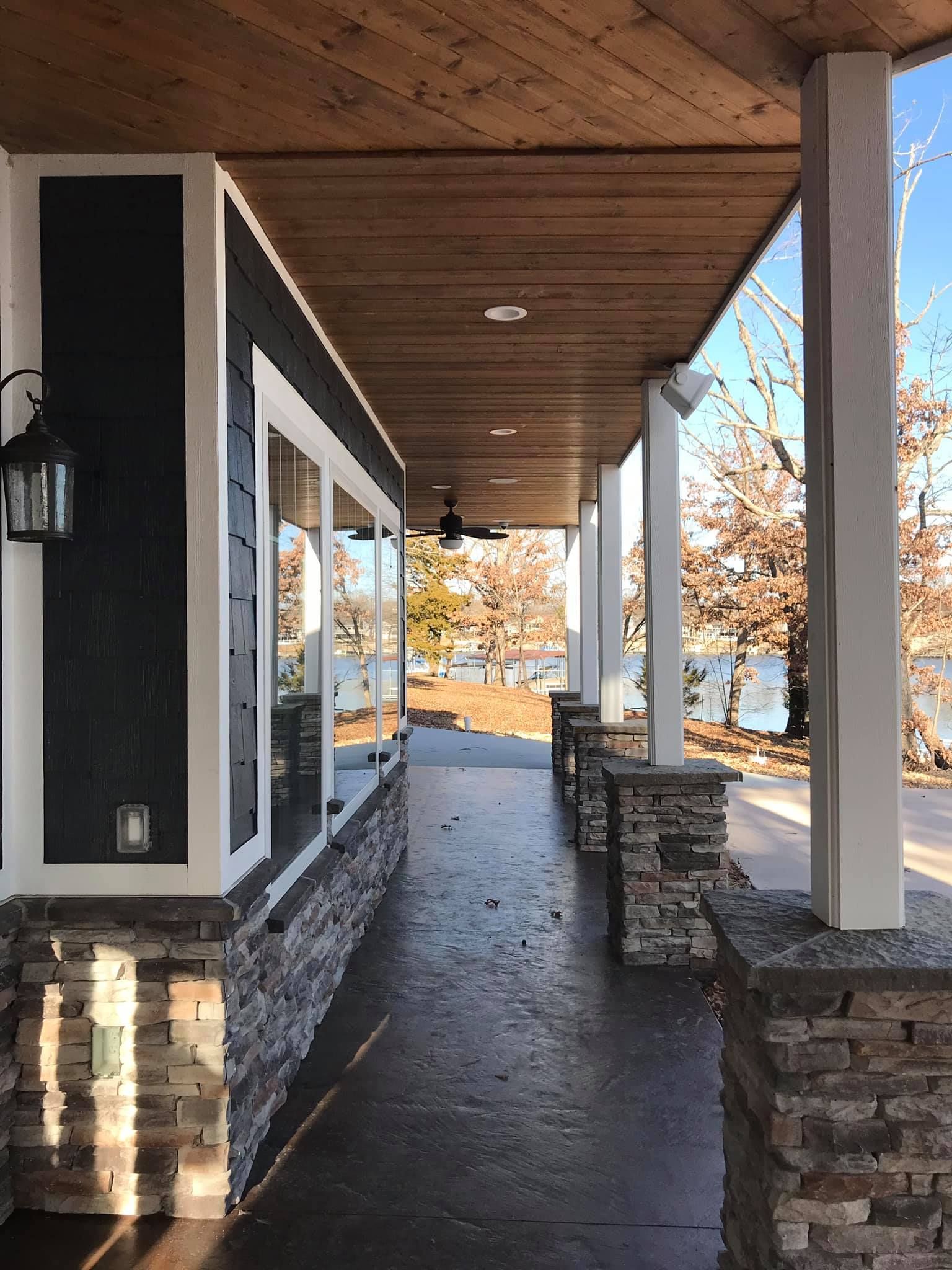 Covered walkway with stone columns, wood ceiling, and large windows beside a building.