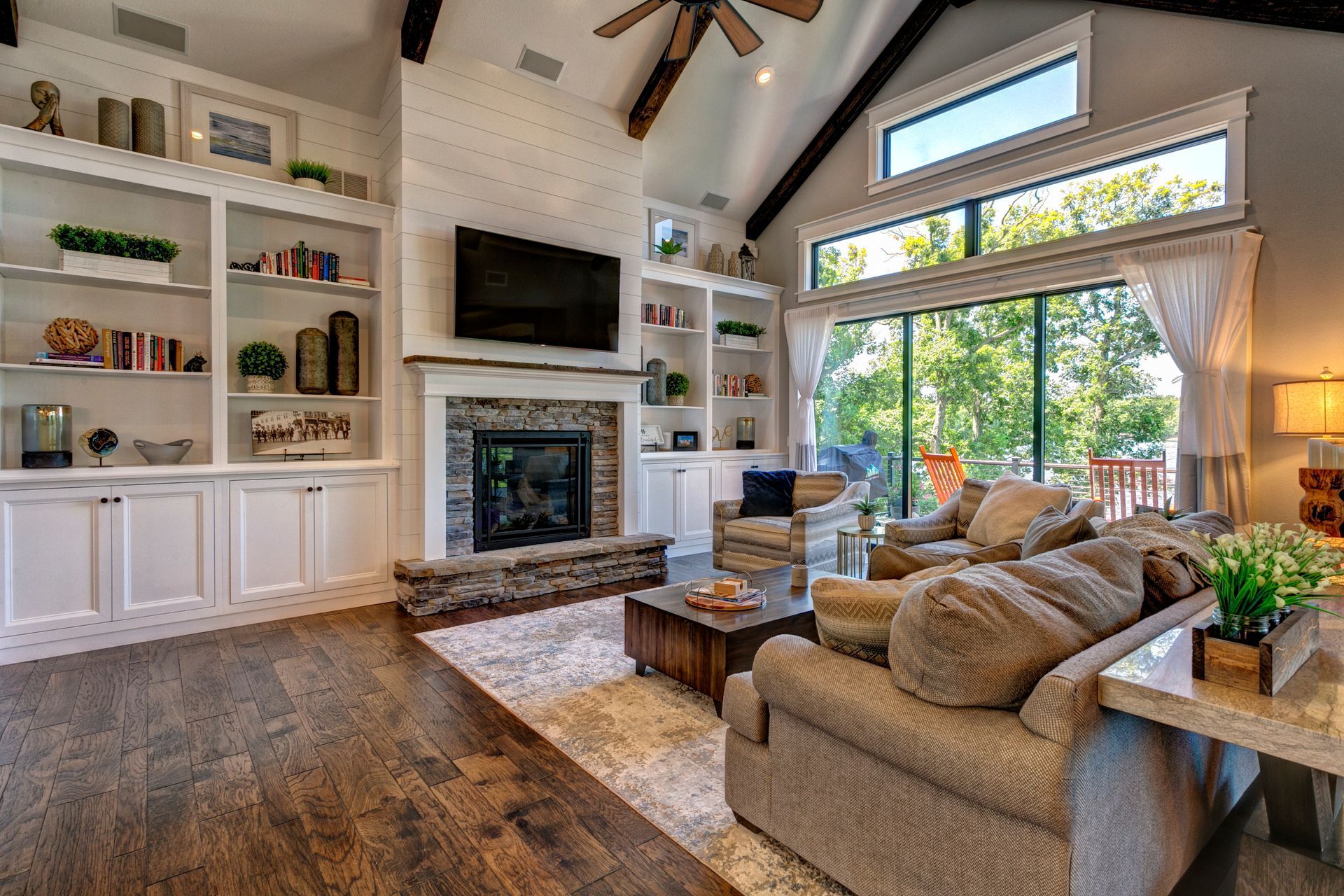 Bright living room with stone fireplace, built-in shelves, tan sofa, and large windows overlooking trees
