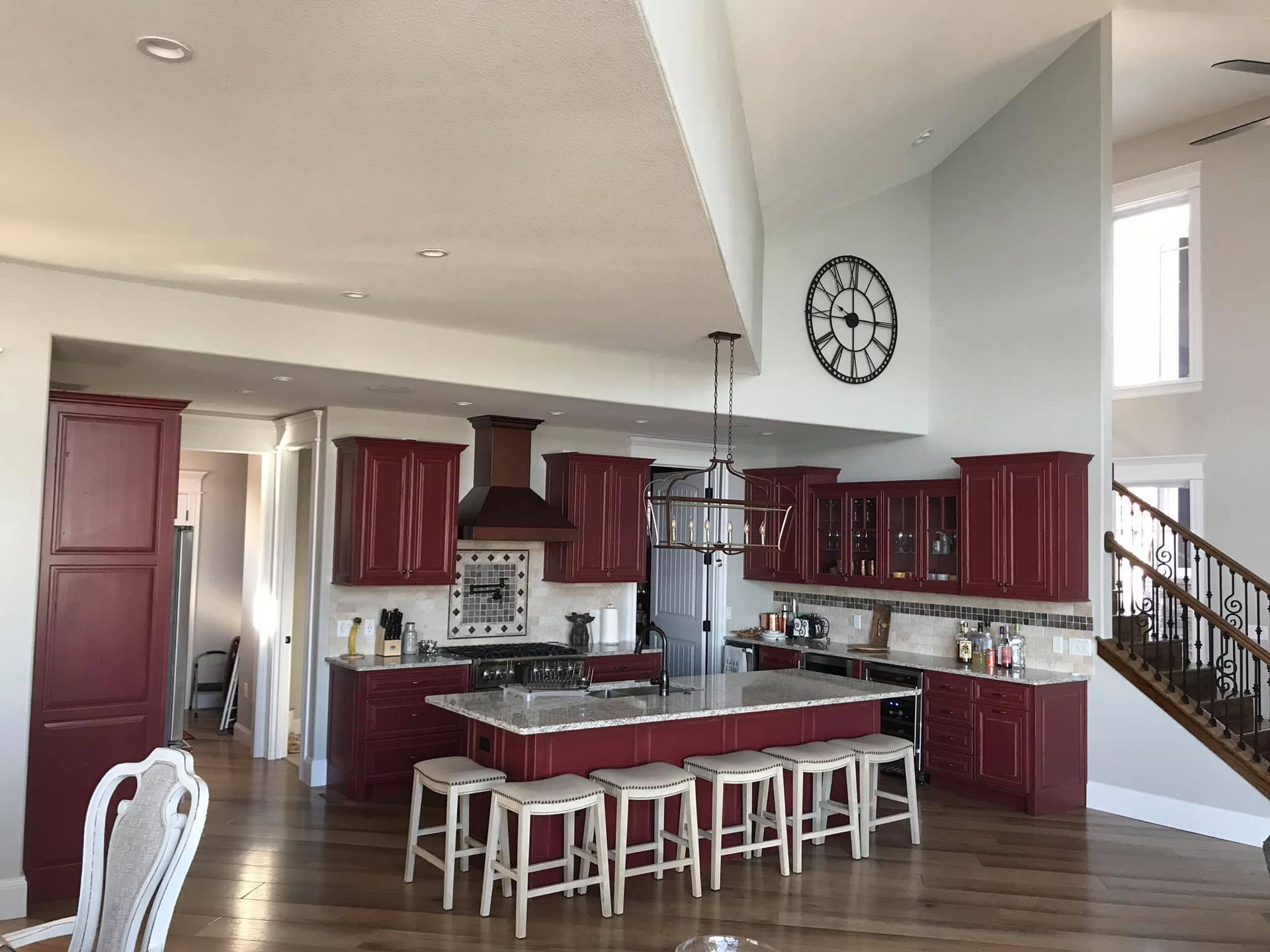 Modern kitchen with burgundy cabinets, island seating, and staircase beside a large wall clock