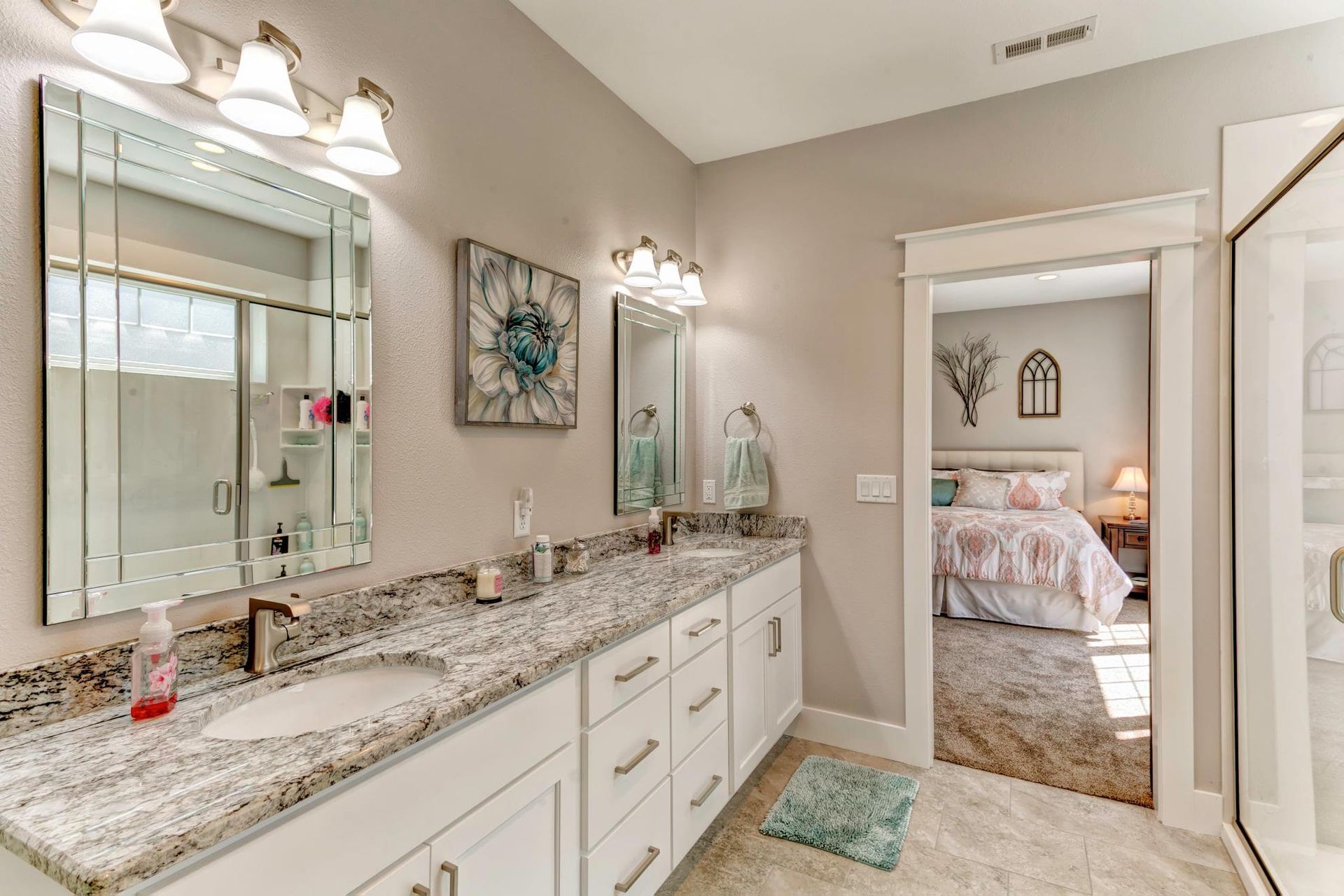 Bright bathroom vanity with double sinks, granite countertop, mirrors, and a glimpse into a bedroom.