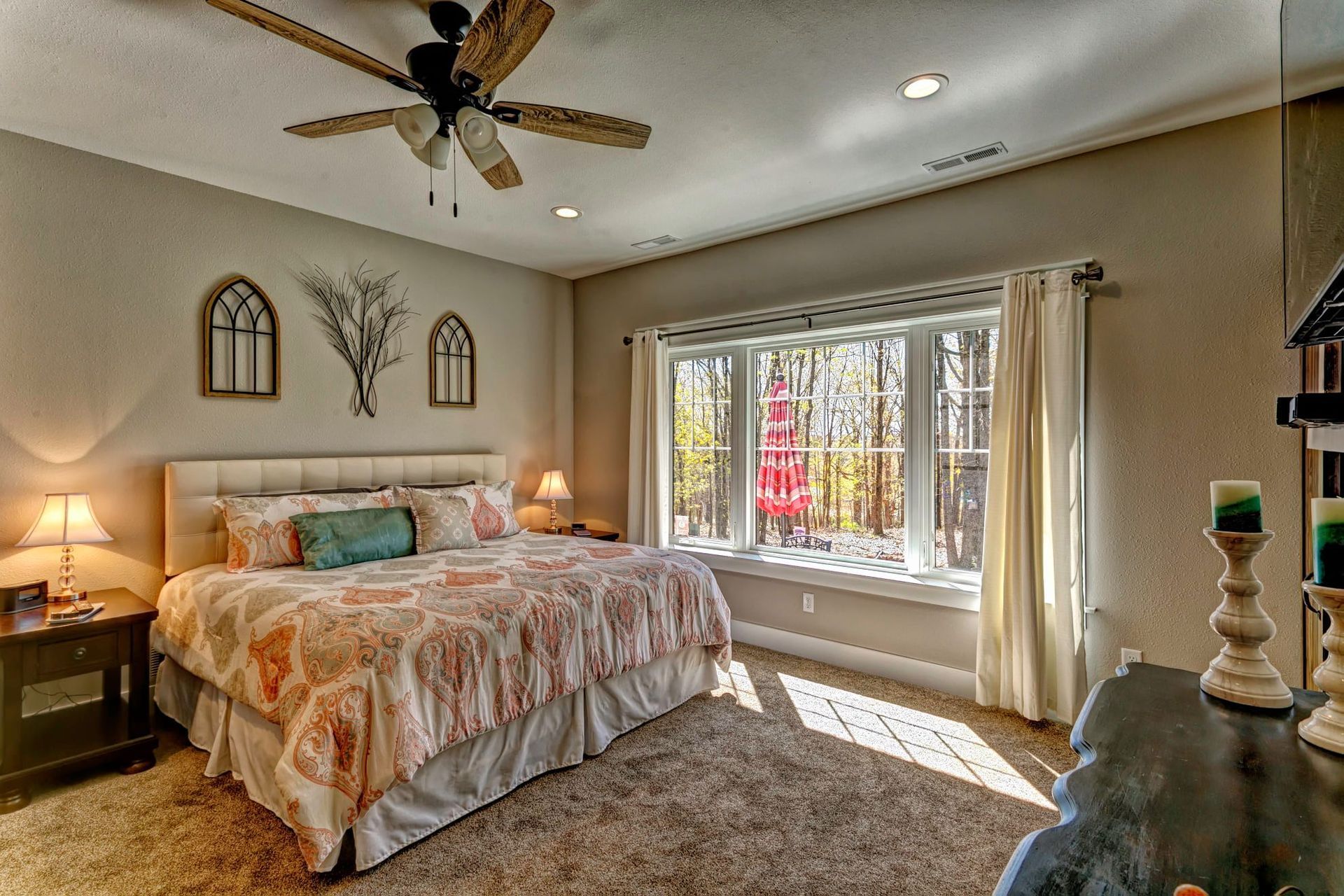 Cozy bedroom with a bed, lamps, ceiling fan, and large window with sunlight and autumn trees outside