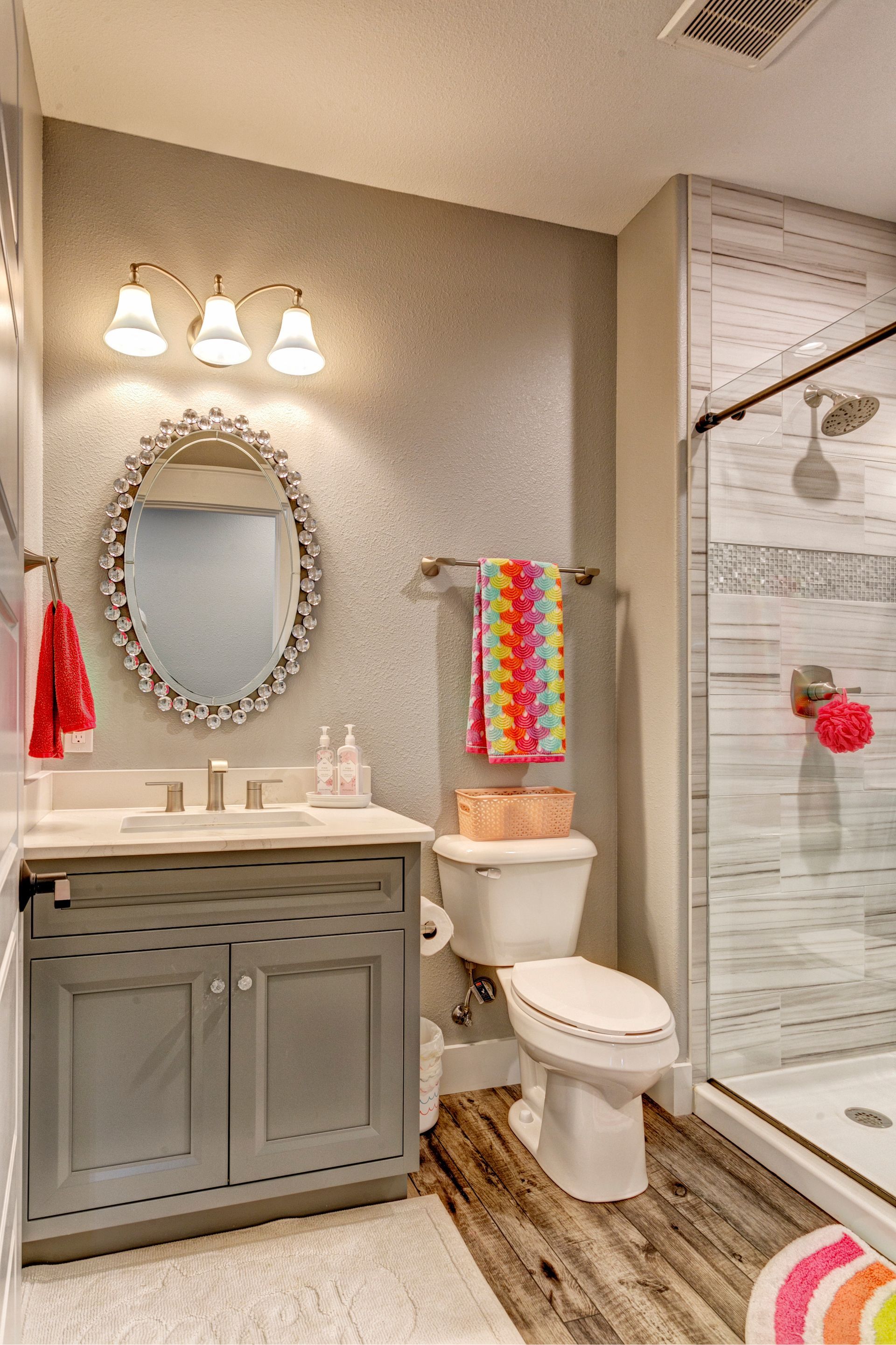 Small bathroom with gray vanity, oval mirror, toilet, and glass shower, accented by red and rainbow towels