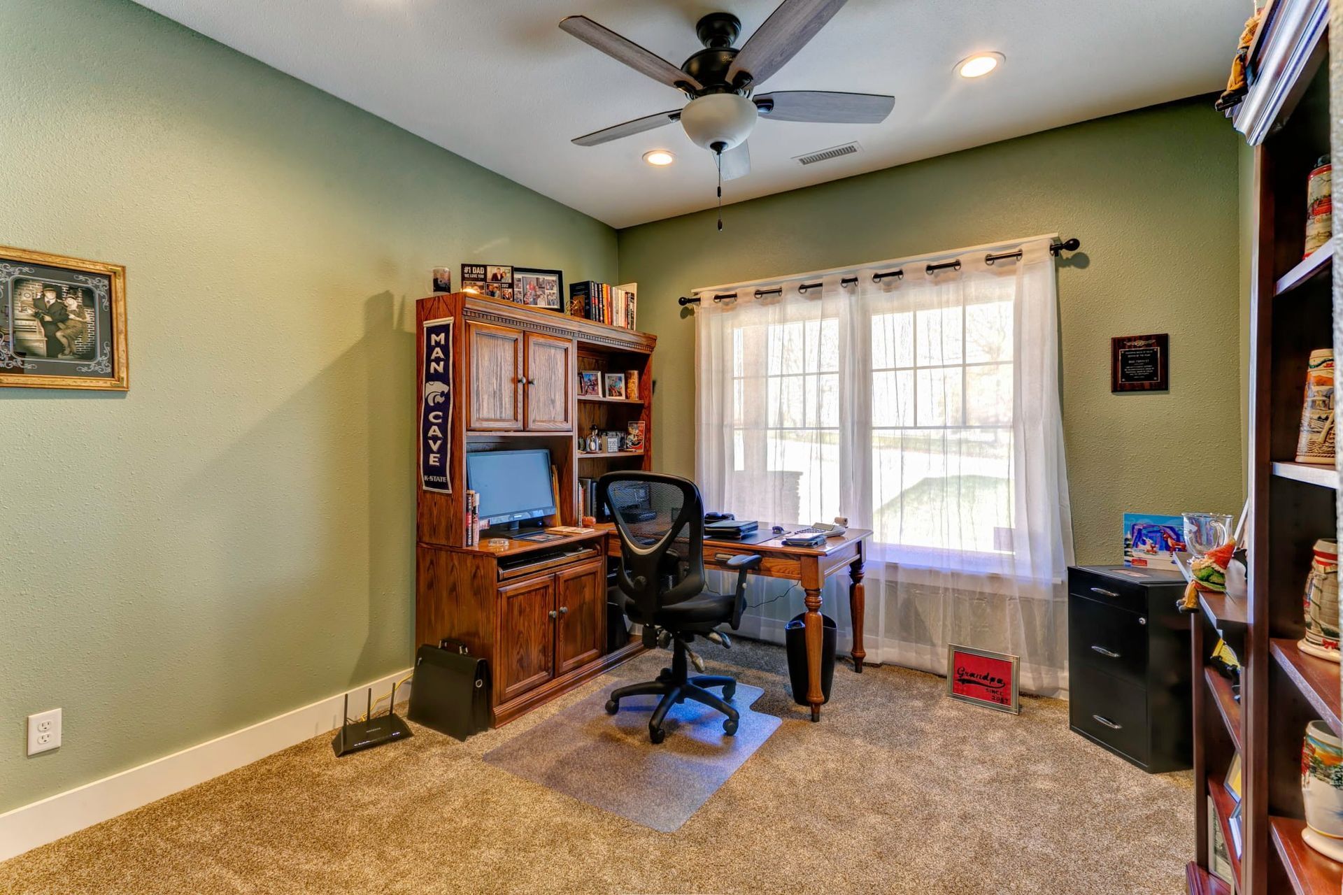 Home office with wooden desk, computer chair, bookshelves, and a large window with curtains