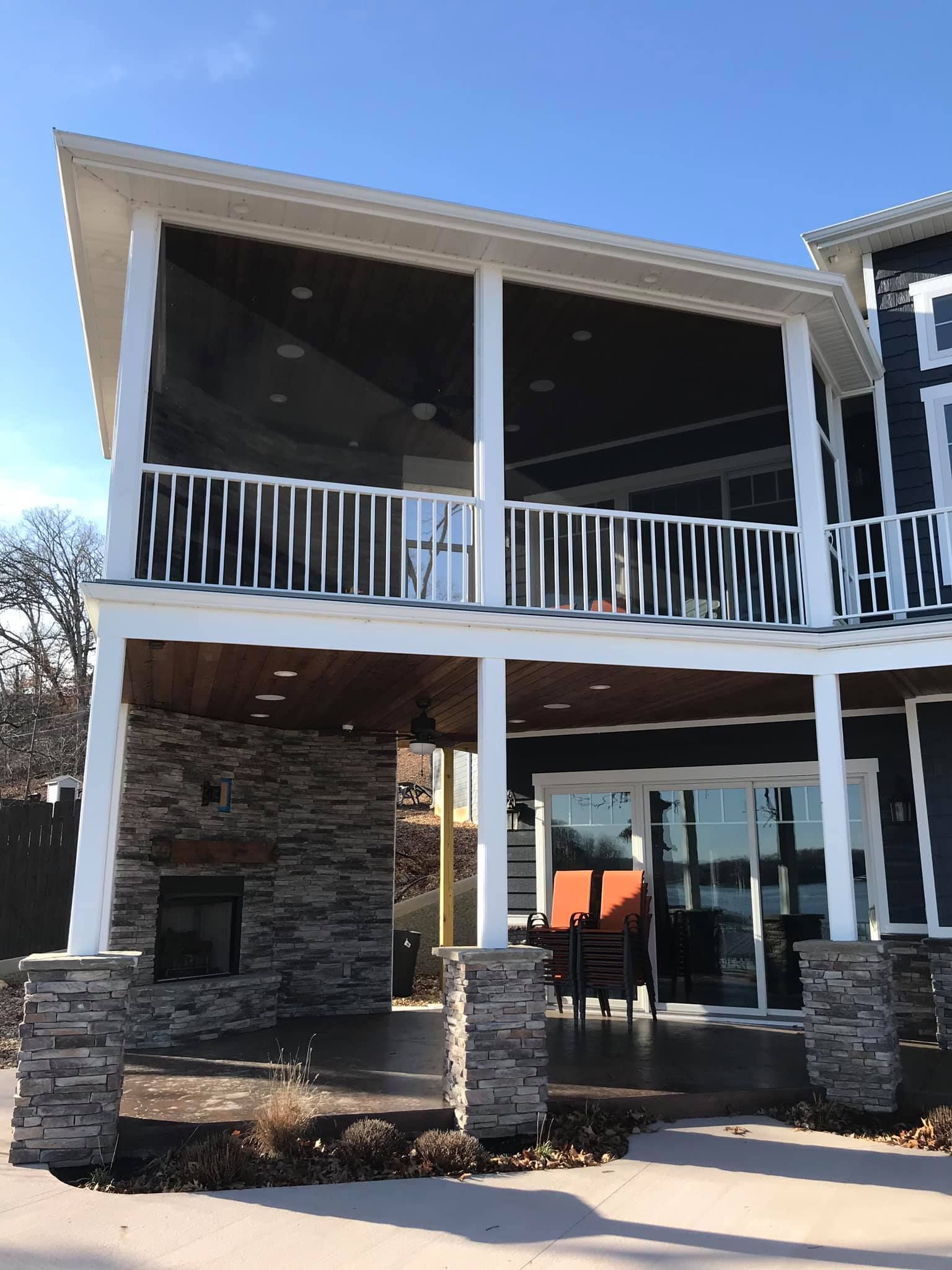 Two-story lakeside house with screened porch, stone columns, and white railings under a blue sky