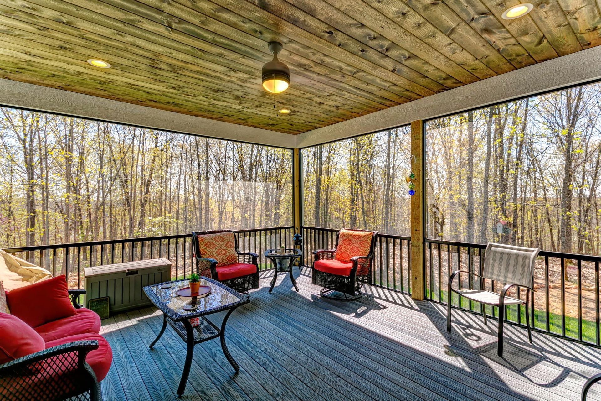 Covered porch with red chairs, rug, and table overlooking a wooded backyard.