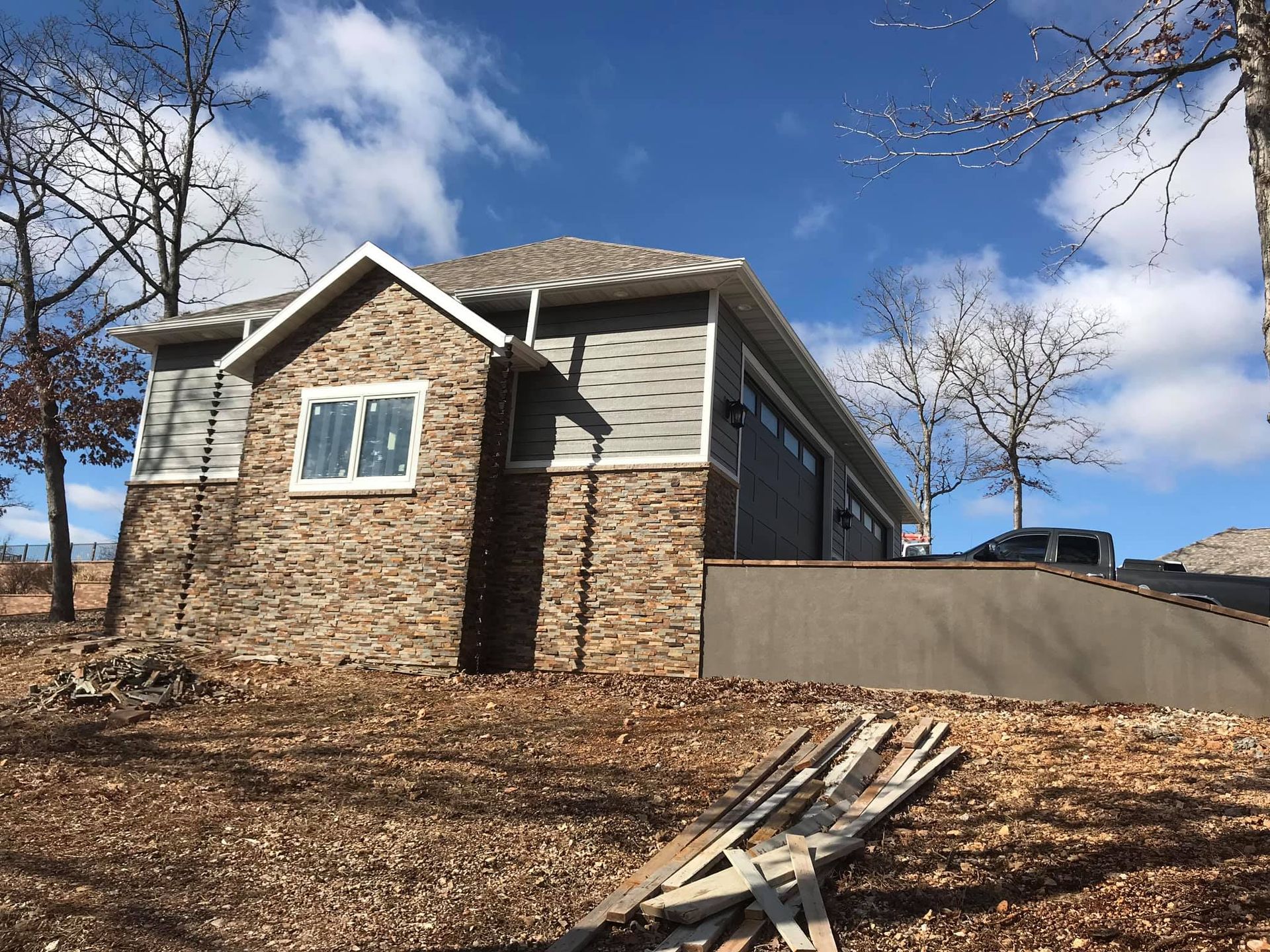 House on a wooded hillside with stone and beige siding, blue sky, and a picnic table in the foreground