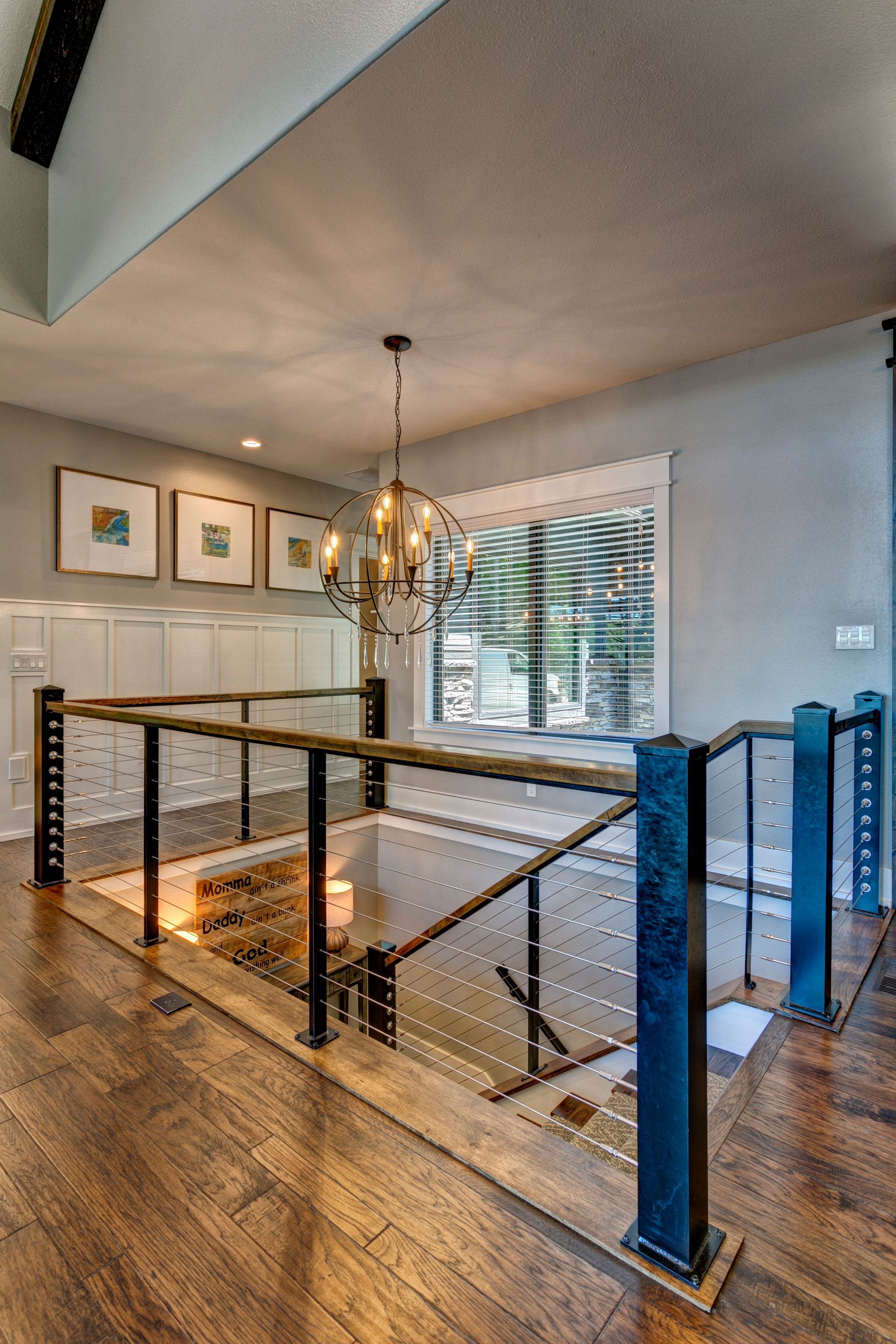 Modern staircase landing with glass railing, wood floors, framed art, and a hanging chandelier￼