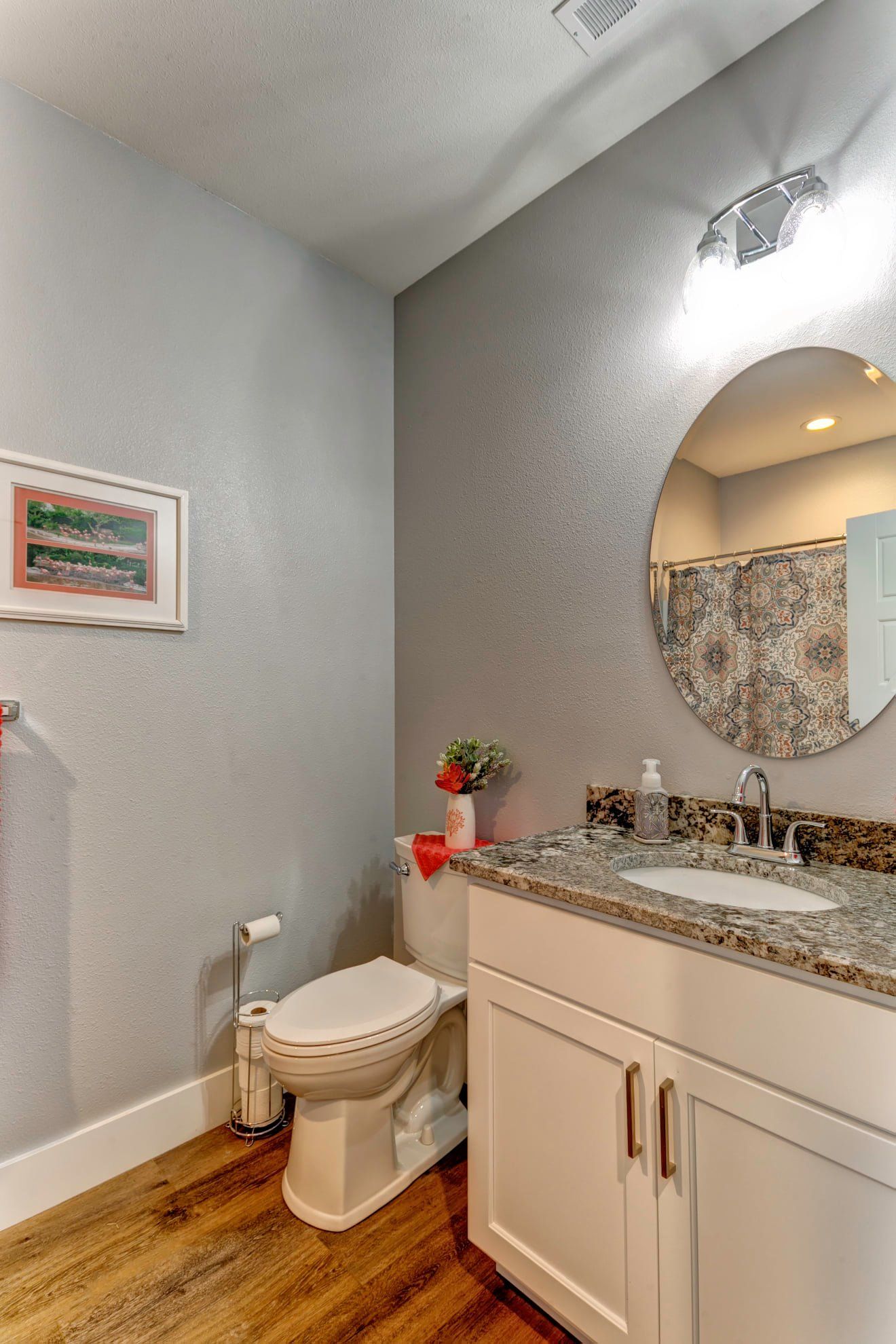 Small bathroom with white vanity, granite countertop, round mirror, and toilet against gray walls.