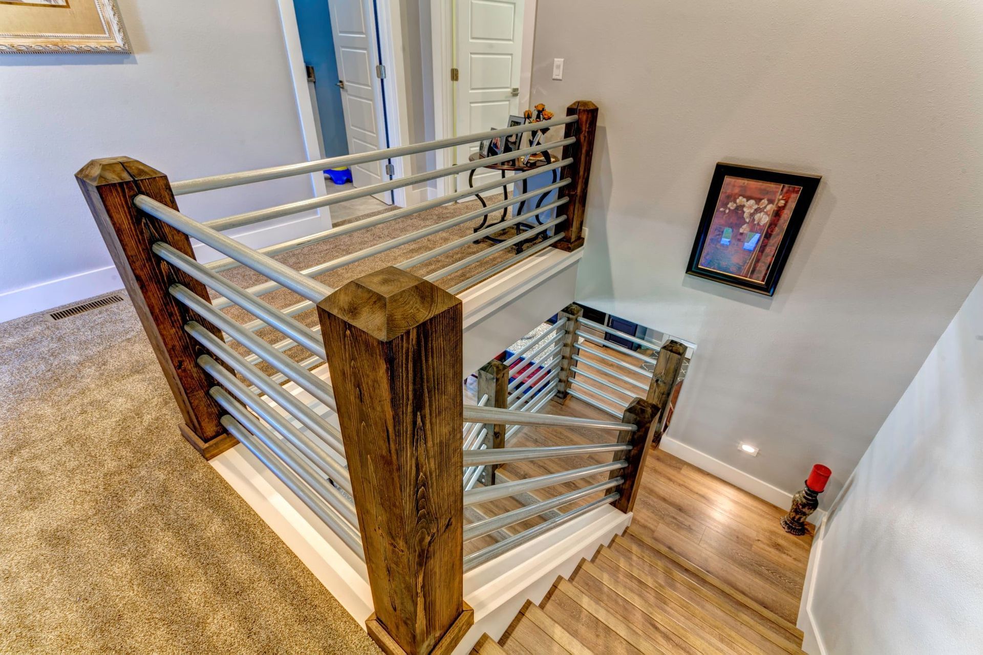 Stairwell with wood posts and metal cable railing overlooking a lower level, with light wood floors and wall art.