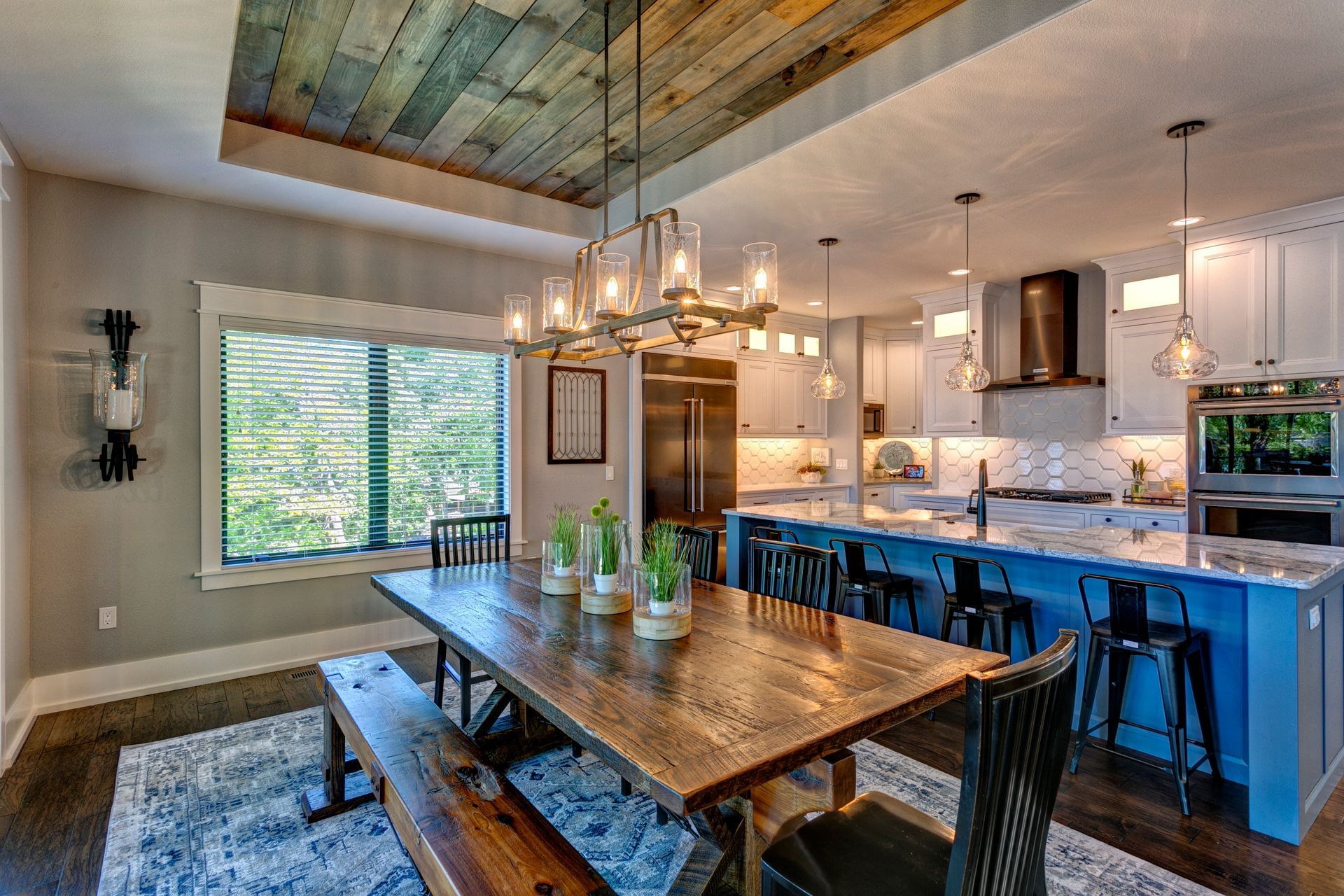 Modern dining room and kitchen with rustic wood table, blue island stools, chandelier, and large window.