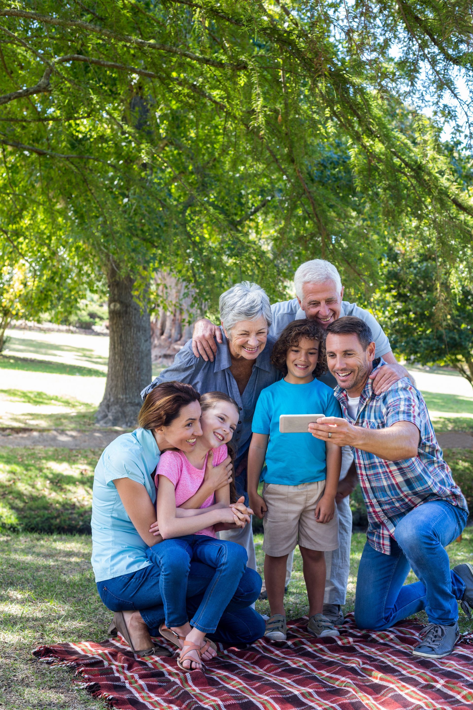 Family group posing together outdoors on a blanket under a leafy tree in a park