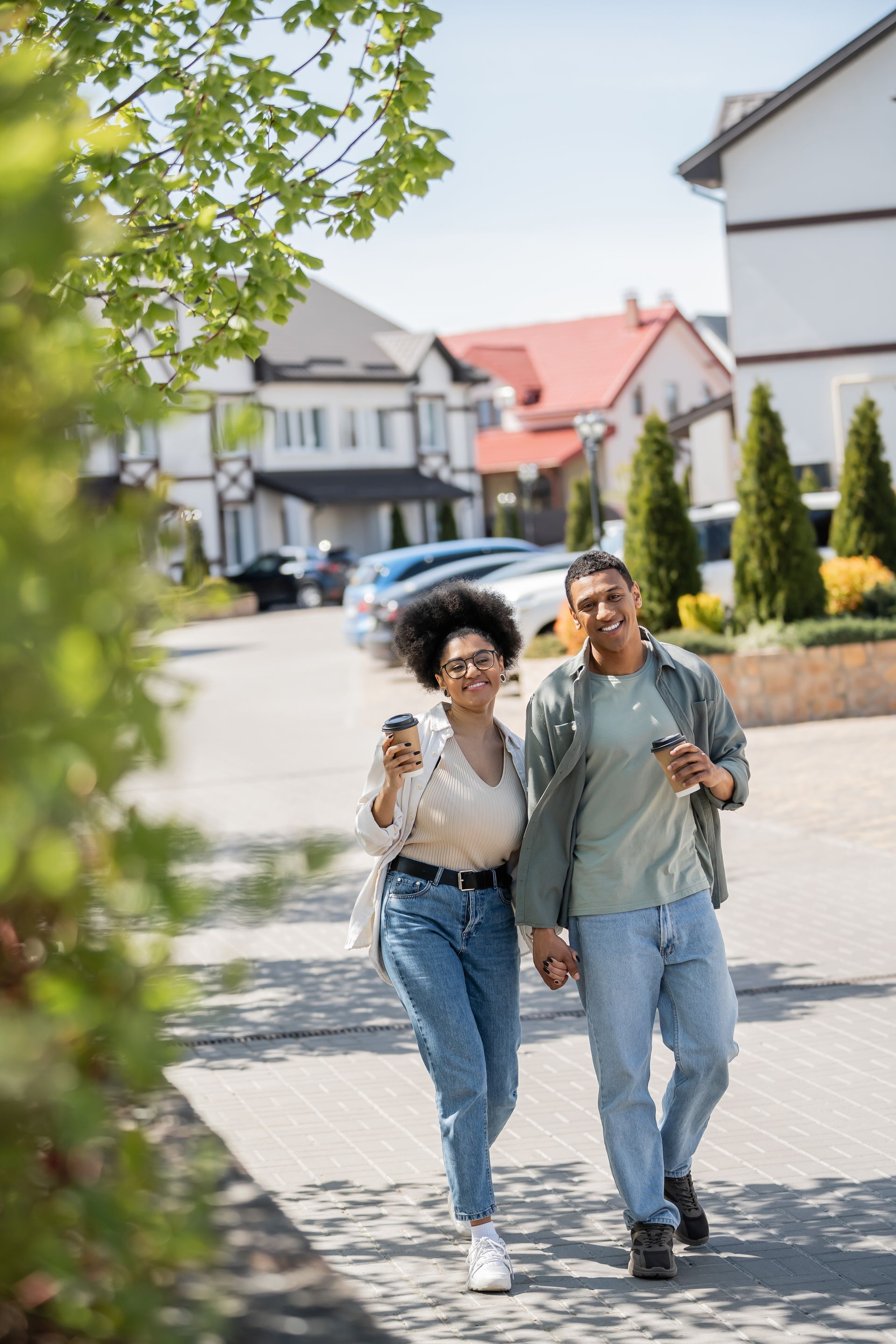 Two people walking arm in arm on a sunny residential street, smiling with houses and greenery behind them.