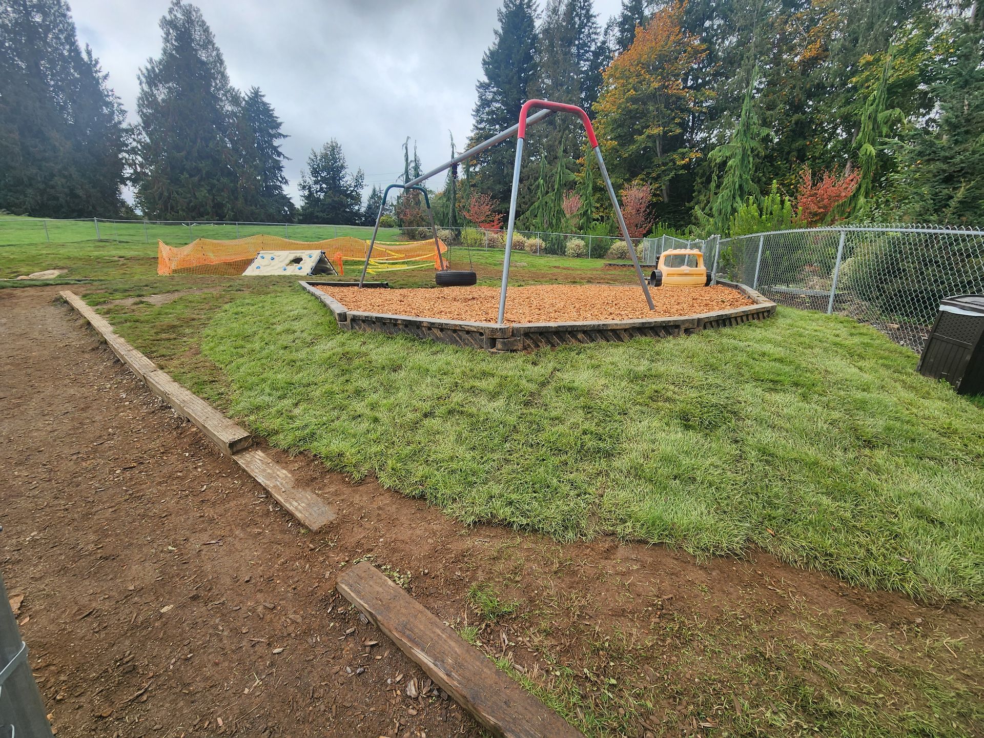 Playground with swings, bark-covered ground, and grassy hill. Gray sky and trees in the background.