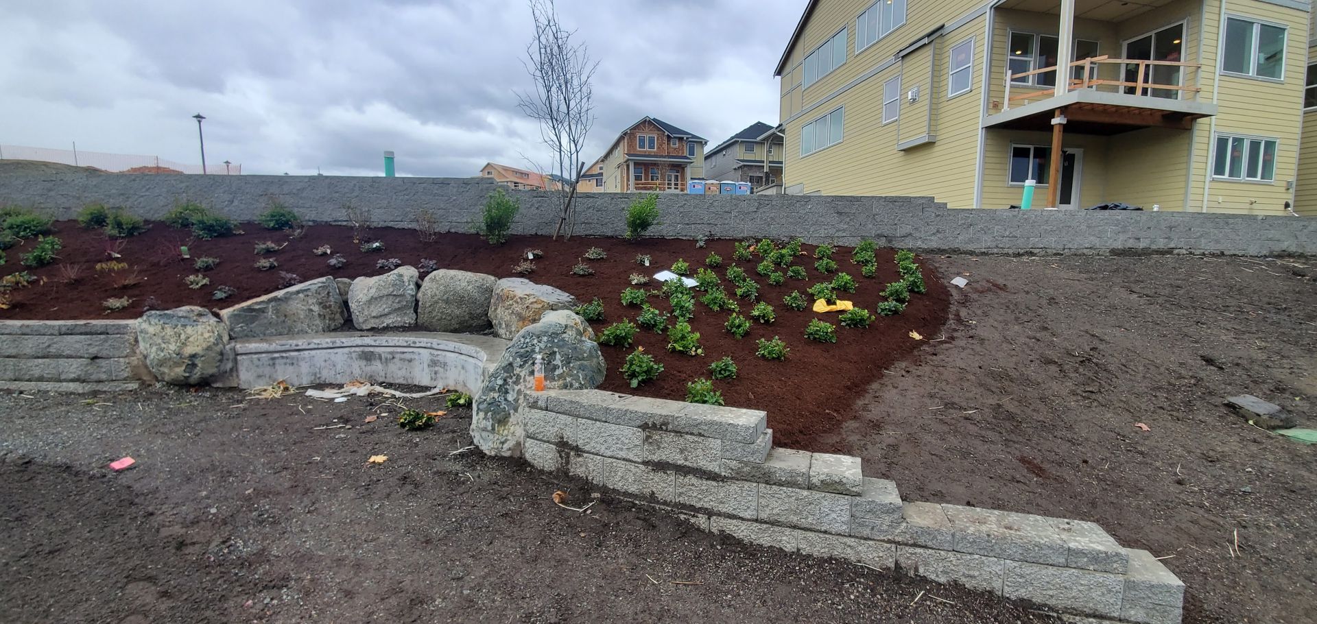 Stone retaining walls and bench in a landscaped yard. Newly planted flowers, brown mulch, and a cloudy sky.