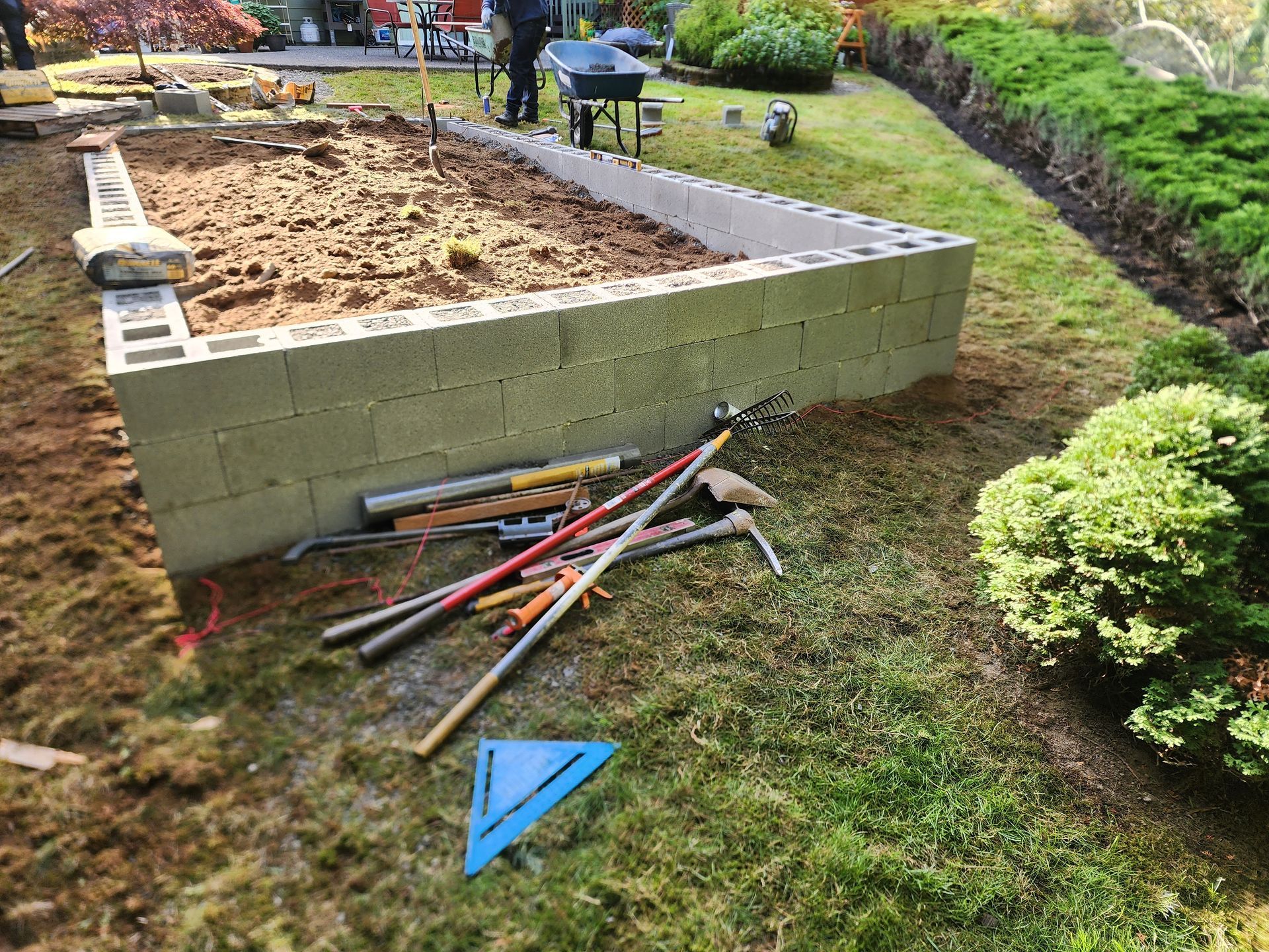 Raised garden bed being built with cinder blocks, tools scattered nearby on grass.