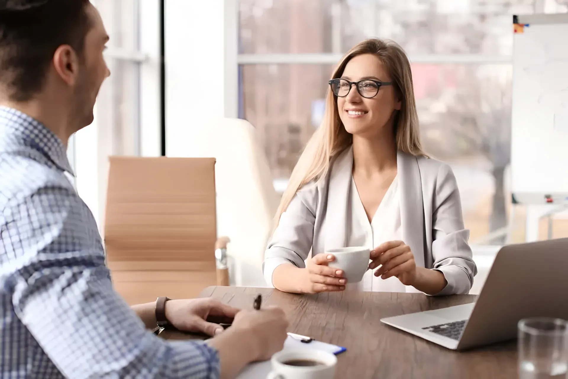 Woman Smiling, Holding a Cup, Interviewing a Man at a Desk — Barolin Physiotherapy Services in Bundaberg South, QLD