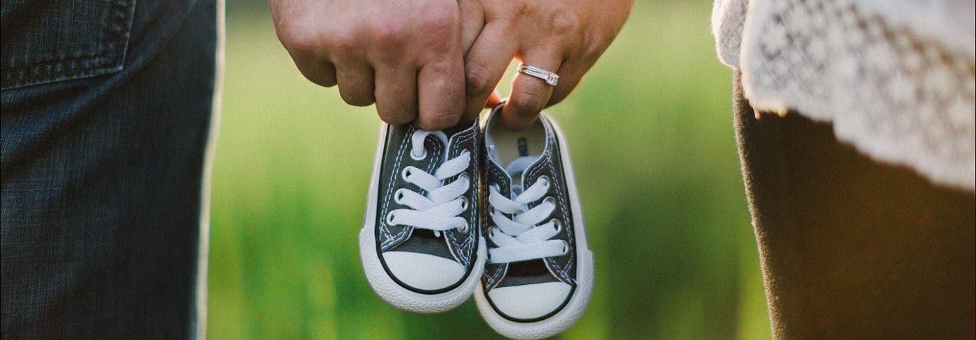 Two hands holding baby shoes, with ring on a finger. Jeans on the left, dress on the right. Green background.