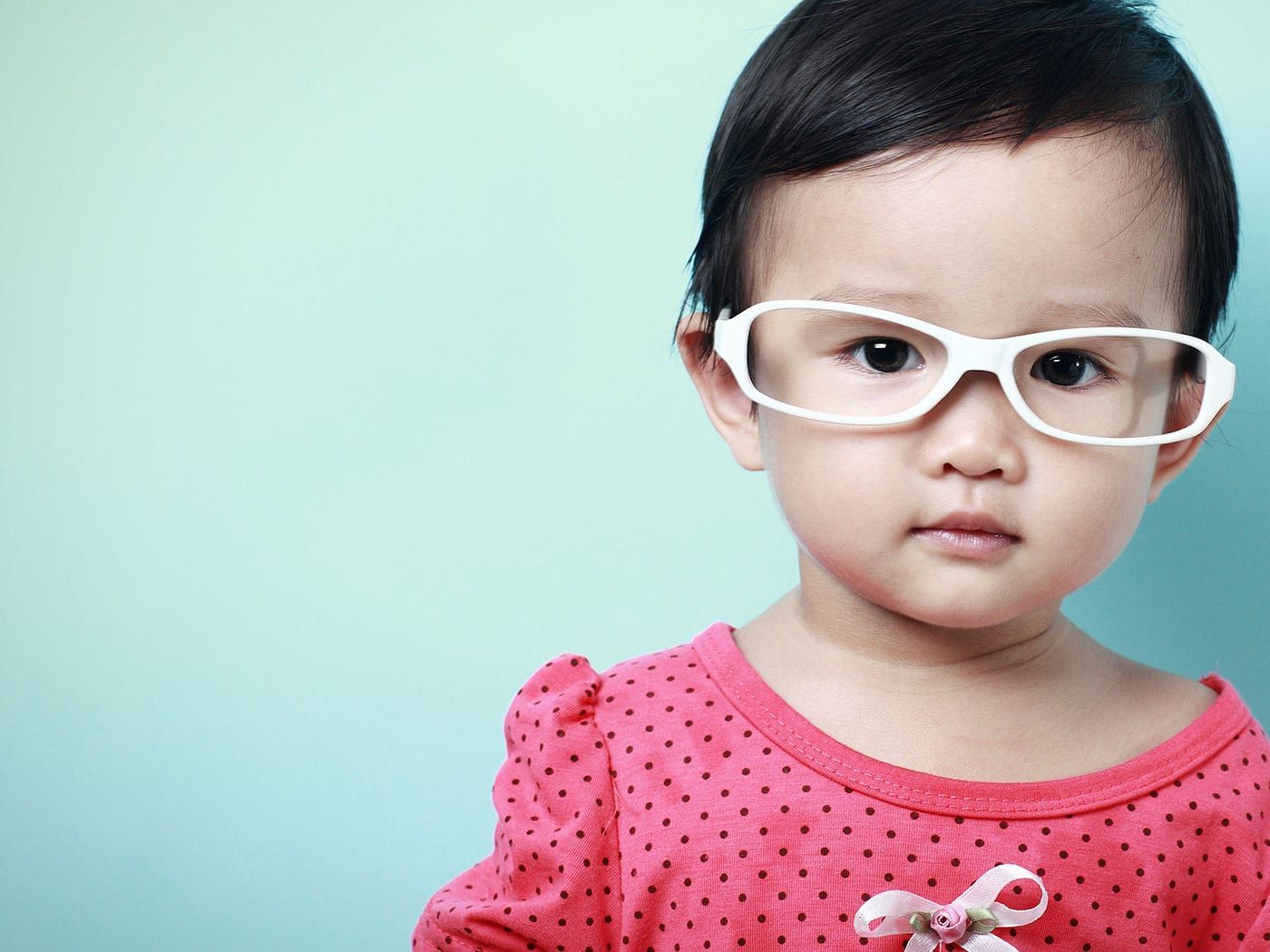 A young child wearing large, white-framed eyeglasses and a polka-dot shirt stands against a light blue background.