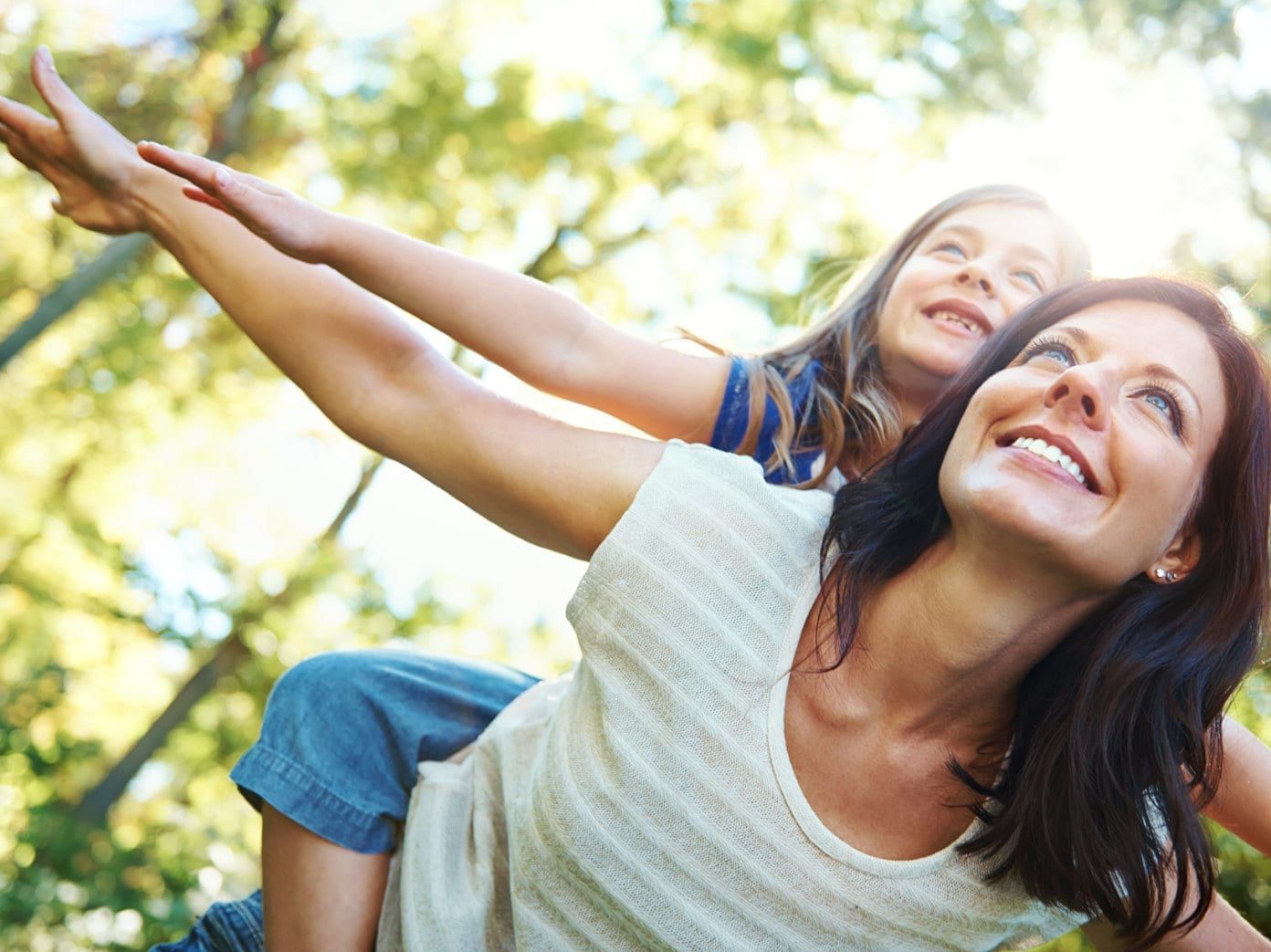 A person carries a child on their back as both stretch their arms out wide and smile in a sunlit outdoor setting.