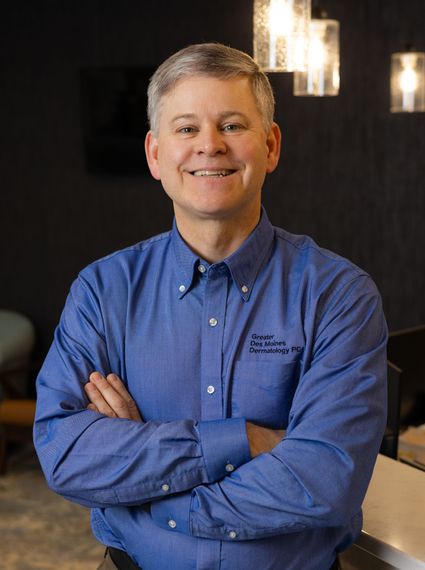 Man in blue shirt, arms crossed, smiling; office setting with lights.