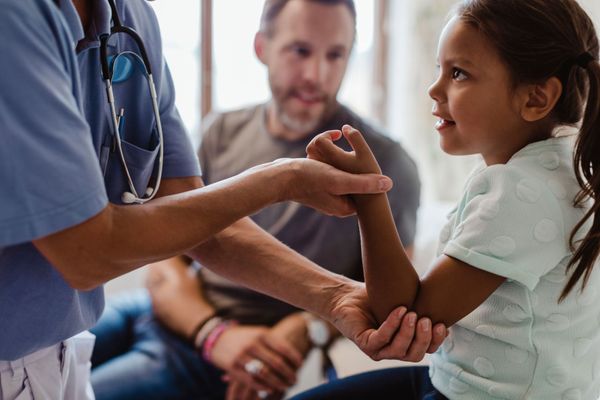 Doctor examining a child's arm in a medical setting, with a parent in the background.