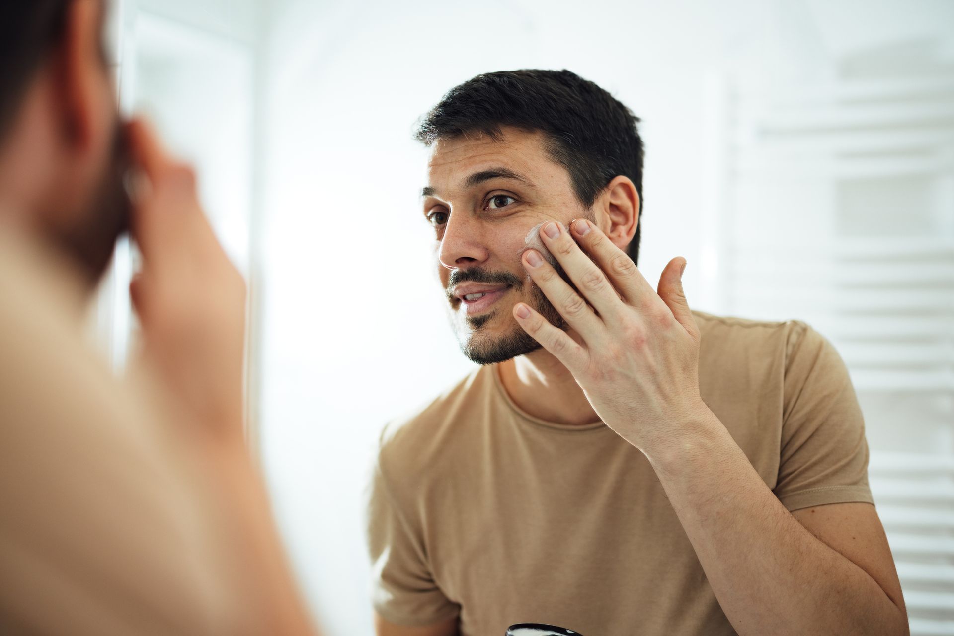 Man applying cream to his face in a bathroom mirror. He wears a beige shirt and has a goatee.