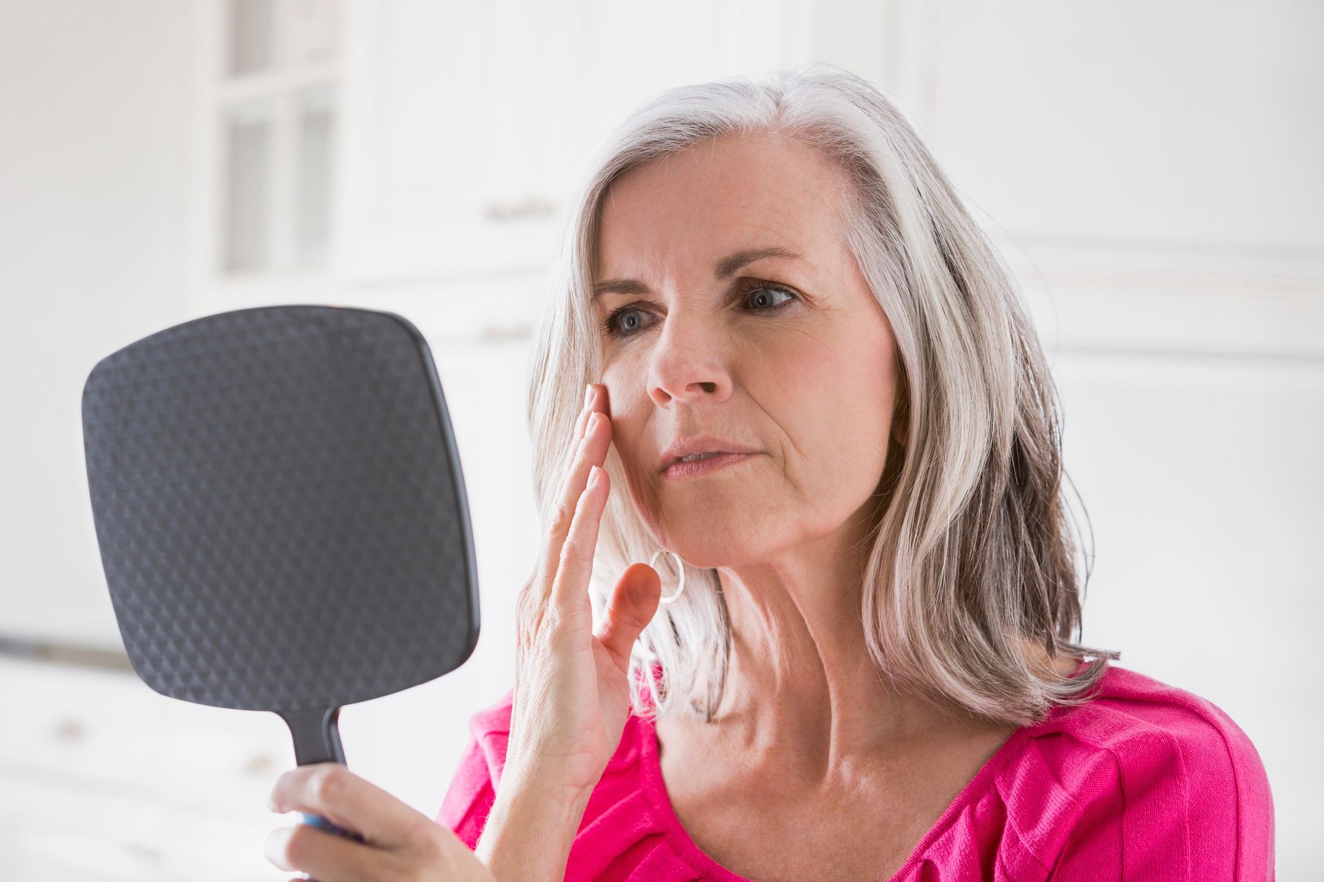 Woman with gray hair examining her face in a hand mirror, indoors. She touches her cheek, looking concerned.