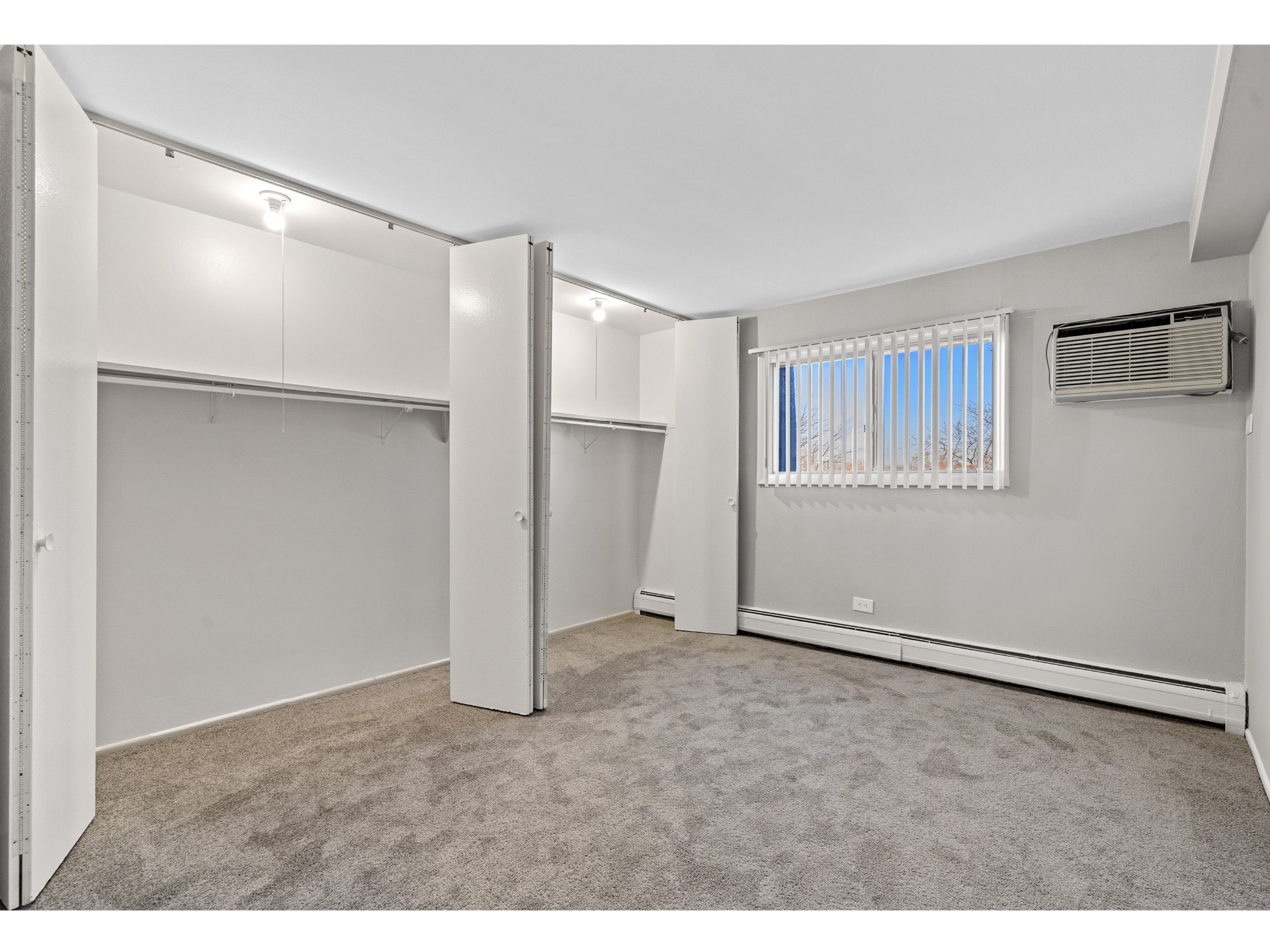 Bedroom with open reach-in closets, beige carpet, window blinds, and a wall AC unit.
