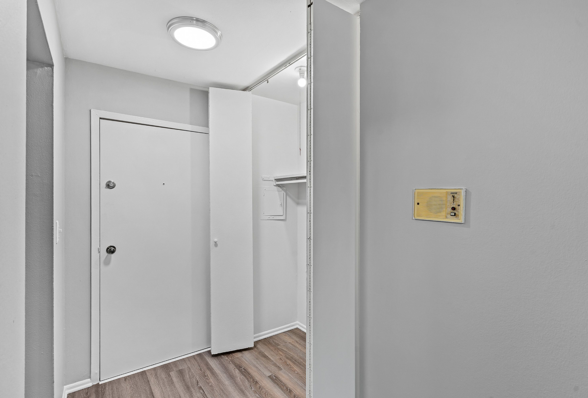 Entryway of an apartment with a white door, open closet, intercom panel, and wood floor.