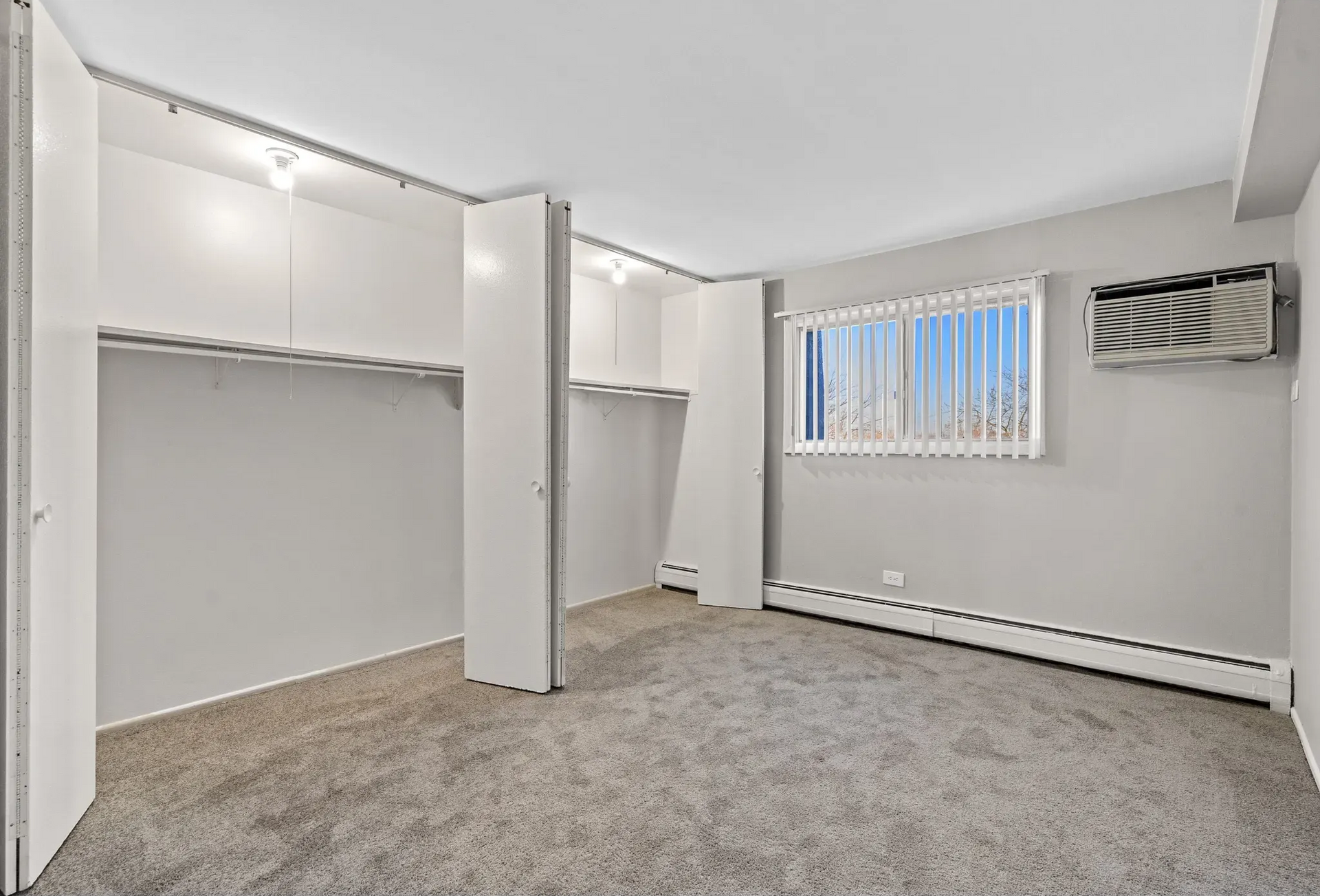 Empty bedroom with built-in white closet. Light gray walls and carpet, window with blinds, and an air conditioner.