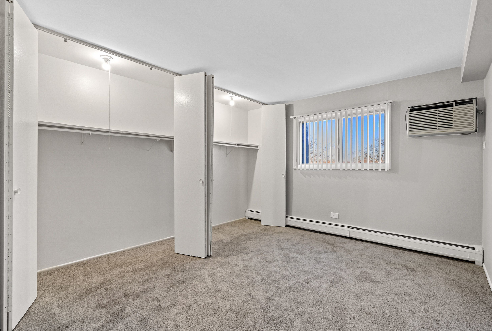 Bedroom with open reach-in closets, beige carpet, window blinds, and a wall AC unit.
