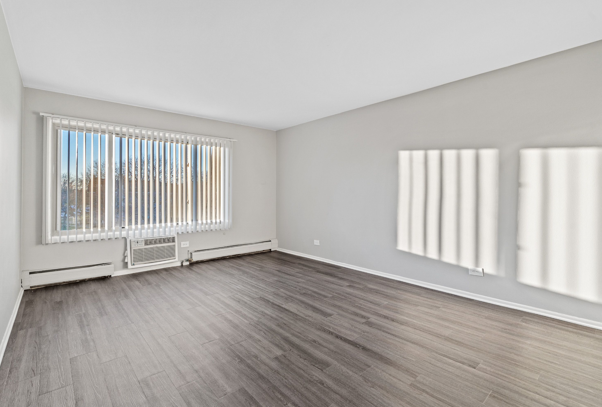 Empty living room with a large window and vertical blinds, gray walls, and wood-look flooring.