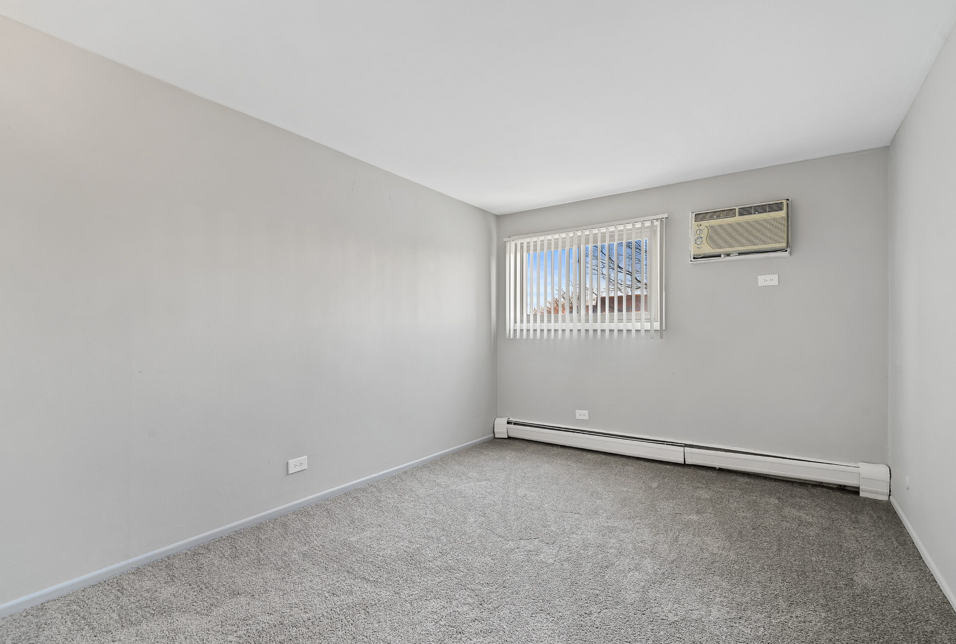 Small empty gray apartment room with a window, vertical blinds, and a wall-mounted air conditioner.
